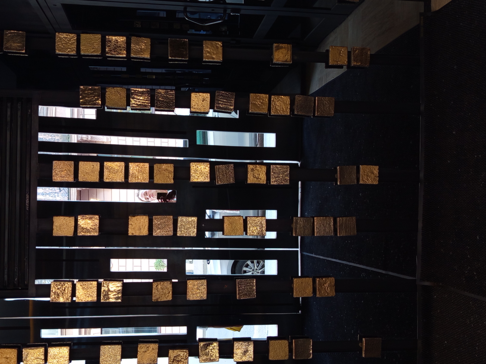 The image depicts a modern entrance with a dark, textured floor and vertical black metal dividers adorned with numerous square, gold-colored decorative elements. These elements, varying in texture and sheen, are arranged in columns at different heights. Through the gaps in the dividers, windows reveal a street scene in Saint Petersburg, Russia, with buildings and a car visible. A person is partially reflected in one of the windows, holding a phone. The overall impression is of an artfully designed entryway, possibly in a commercial or residential building, with a view of the urban surroundings.