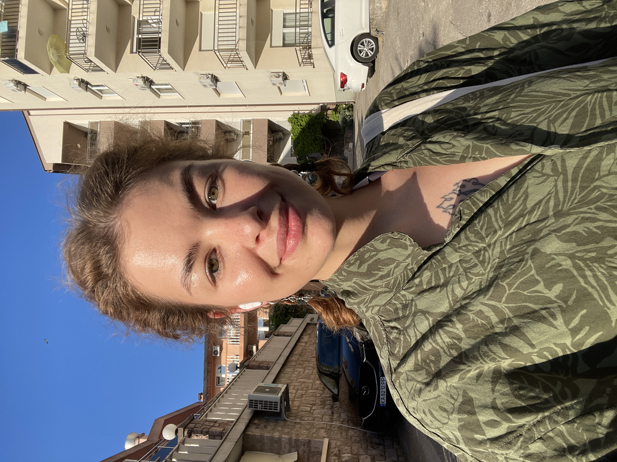 A woman with fair skin, green eyes, and light brown hair partially pulled back takes a selfie in Rafailovići, Montenegro. She is wearing a green shirt with a leaf pattern and a white strap is visible over her shoulder. She is standing on a street lined with buildings, including an apartment building with white balconies, and a car with license plate "KA 8212 PP" is parked nearby. The sky is blue, and the sun is shining.