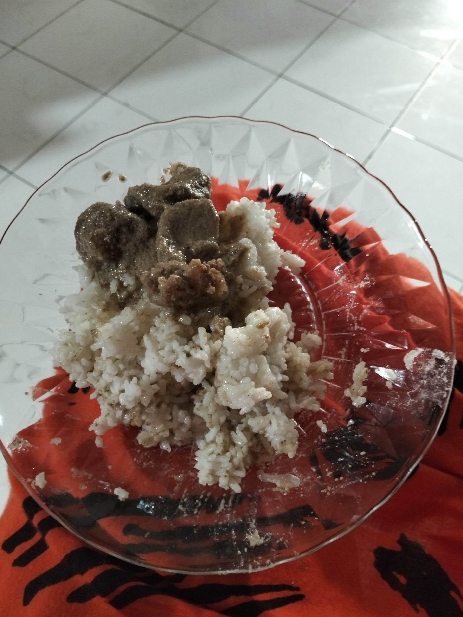 A close-up shot shows a clear glass plate filled with white rice and cubed meat. The rice is piled high on the left side of the plate, while the meat, coated in a dark gravy, is placed on top of the rice and also to its right. The plate is set on an orange cloth with a black striped pattern. The background is a tiled floor with white squares and gray grout lines, illuminated by overhead lighting. The lighting creates a bright, reflective glare on the tiled surface, suggesting an indoor setting. No people are visible in the image. The food appears to be a meal, likely served in a home environment in Payagaboh, Indonesia, given the location context. The scene is well-lit, and the focus is entirely on the food and the plate. There is no visible text in the image.