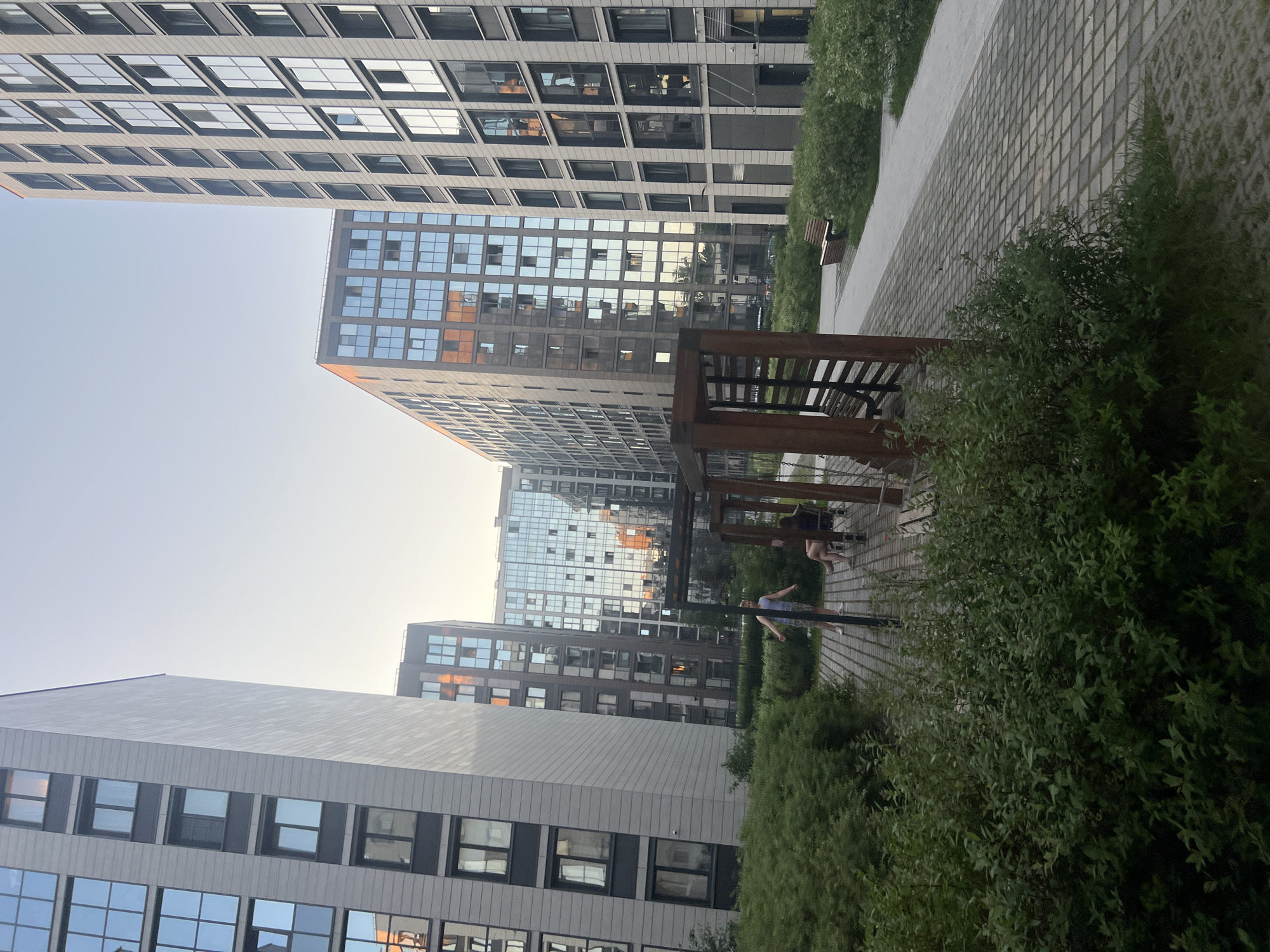 The image depicts an outdoor scene within a modern residential complex in an unknown city. Multiple high-rise buildings with contemporary architecture define the background and midground, bathed in the soft, warm light of late afternoon or early evening, indicated by a pale blue sky transitioning to a yellowish hue at the horizon.
On the far left, a tall, multi-story building with a light grey facade and dark grey window framing features numerous rectangular windows, many reflecting the sky. Towards the center and right, other multi-story buildings display reflective glass facades, with some windows exhibiting distinct orange and reddish-brown reflections from the direct sunlight. The building on the far right appears to have a slightly darker facade and larger windows on its ground level.
In the midground, a paved area with square grey pavers forms a pathway or communal space. Dense green bushes and foliage line this path, particularly prominent in the foreground and along the left side. Centrally located on the paved area is a wooden structure, possibly a pergola or covered seating area, featuring thick wooden posts and at least two attached wooden benches.
At least three individuals are present near this structure. A light-skinned male, wearing a light sleeveless top and patterned shorts, stands upright with arms slightly spread, positioned to the left of the wooden structure. Two light-skinned females are seated on one of the wooden benches within the structure. One female, with dark hair, wears a dark top and shorts, with her legs crossed at the ankles. The other female is partially obscured, visible from the waist down, seated next to the first on the same bench. Their interaction suggests a casual social gathering or relaxation. A third wooden bench is visible further along a paved path in the background, near more green landscaping.