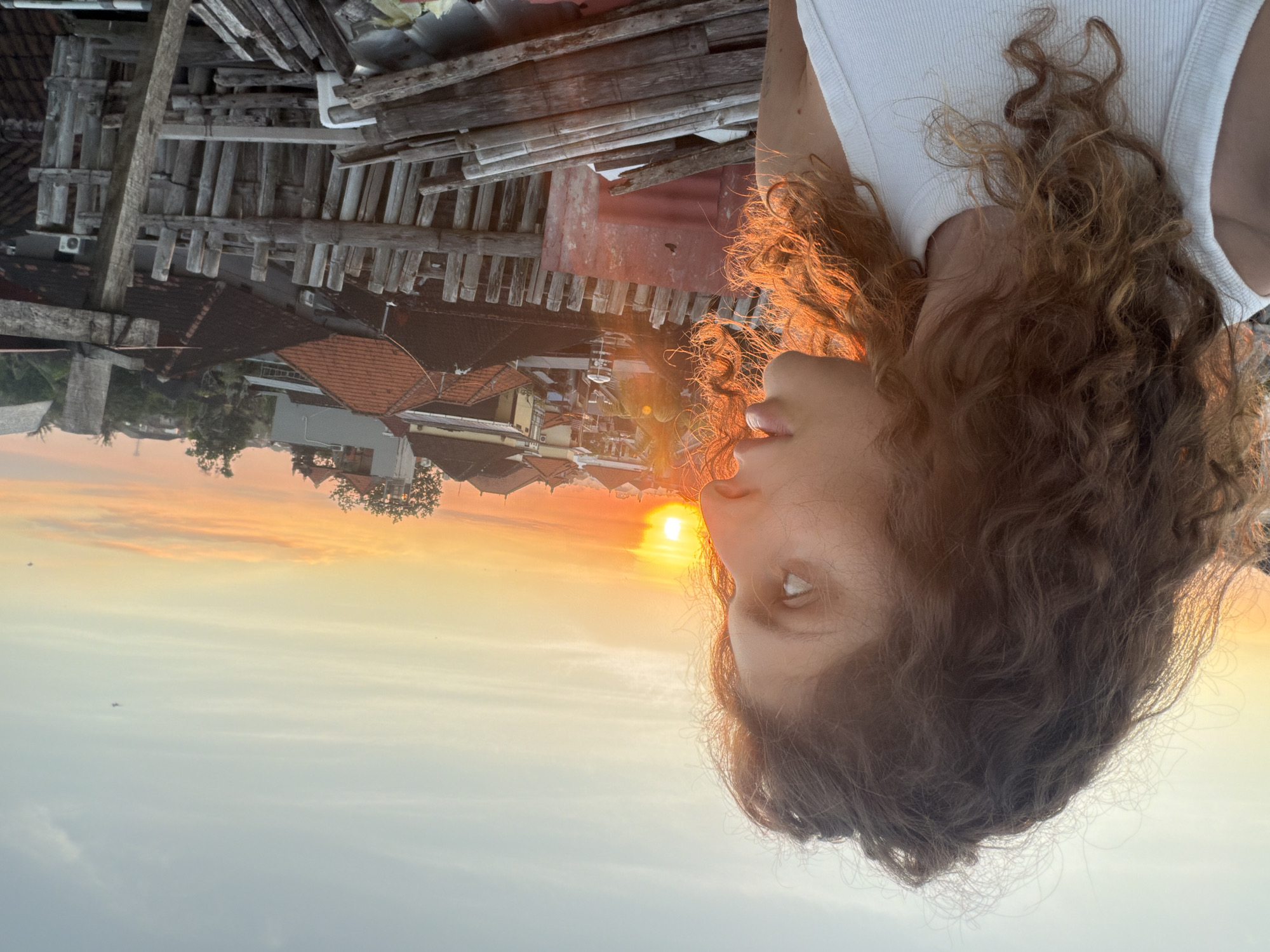 The image depicts a person with curly brown hair and fair skin wearing a white tank top. The person's head is partially visible, facing towards the left of the frame. The background includes buildings with red tile roofs, suggesting a residential area. Above the buildings, there is a clear sky with colors ranging from light blue to orange, indicating a sunset. In the top portion of the image, there is what appears to be a wooden structure. Given the location of North Kuta, Indonesia, it's likely this is a rooftop or an elevated structure with a view of the surrounding buildings and sky at sunset.