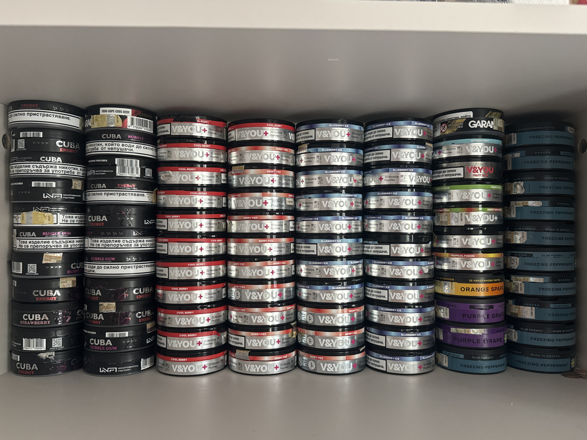 This media file shows a tightly packed shelf filled with various brands and flavors of nicotine pouches. The items are arranged in neat stacks, forming columns and rows. The dominant colors are black, white, red, blue, and purple, corresponding to the packaging of the pouches.
The brands clearly visible include "CUBA" in various flavors like Energy, Bubble Gum, and Strawberry, and "V&YOU+" in flavors such as Cool Berry, Berry Fizz, and Blueberry Ice. Other brands like "GARAN" and "FREEZING PEPPERMINT" are also present. Many of the pouches are labeled in Bulgarian, with phrases such as "Това изделие съдържа никотин, който води до силно пристрастяване" (This product contains nicotine, which leads to strong addiction) and "Не се препоръчва за употреба от непушачи" (Not recommended for use by non-smokers). Other text visible includes flavor names, nicotine content (e.g., "20 Nicotine Pouches"), and origin information like "BORN IN SWEDEN".
The setting appears to be a retail shelf, possibly in a store or a personal collection. The lighting suggests an indoor environment, likely under artificial light. There are no people or animals visible, and the focus is solely on the arrangement of the nicotine pouch products. The overall impression is one of a well-stocked display of these specific consumer goods.