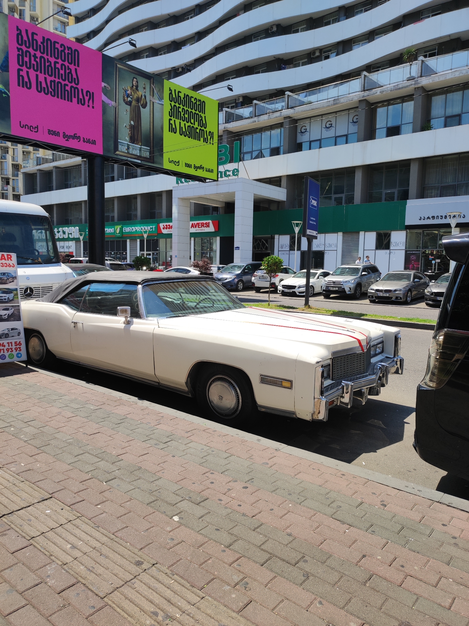 In Batumi, Georgia, on a sunny day, a white convertible Cadillac Eldorado from the 1970s is parked on a sidewalk. The car features a black convertible top and red stripes on its hood. It is positioned next to a black SUV, partially obscuring the view of the Cadillac's rear. The sidewalk is paved with reddish-brown and grey bricks. In the background, a modern white building with curved balconies and large windows rises high, featuring shops on the ground floor. Among these businesses are "EUROPRODUCT", "AVVERSI APOTHEKA", and signs with Georgian text. Billboards with neon pink and green backgrounds display Georgian text and imagery, including a painting of a woman holding objects. A white bus is also visible on the street. The scene captures a street view in a modern urban environment with a prominent display of vintage American automotive design.