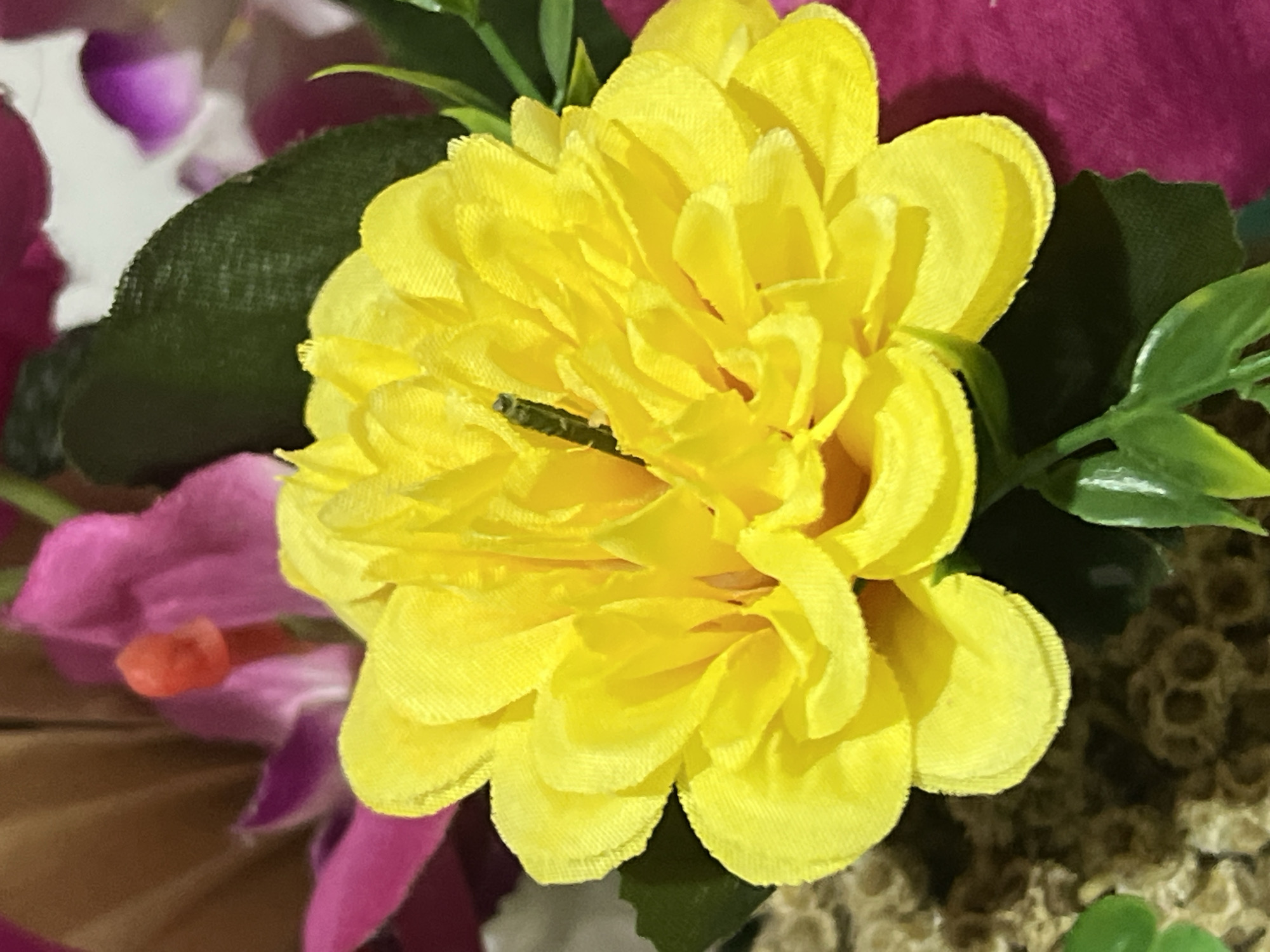 A close-up, eye-level shot focuses on a bright yellow, artificial flower, likely part of a larger floral arrangement. The flower is multi-petaled and vibrant, with a darker green stamen visible at its center. Behind the yellow flower, parts of other artificial flowers are visible, including a deep pink one with a reddish-orange stamen, and hints of purple and white. Dark green artificial leaves are interspersed throughout the arrangement. The background is a blur of colors, suggesting a shallow depth of field. The scene is likely indoors, given the artificial nature of the flowers. It's not possible to determine the specific location in Denpasar, Indonesia, from the image alone.