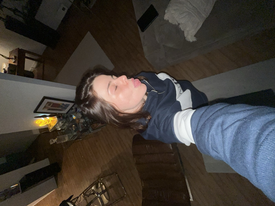 A woman with brown hair, wearing a dark blue and white sweatshirt and a white shirt underneath, is lying on a wooden floor. Her eyes are closed, and her tongue is sticking out slightly. A couch is visible in the background, along with a black phone and a white throw. To the left, a glass table and a picture frame sit on a shelf. A vintage standing lamp is also visible in the lower left corner.