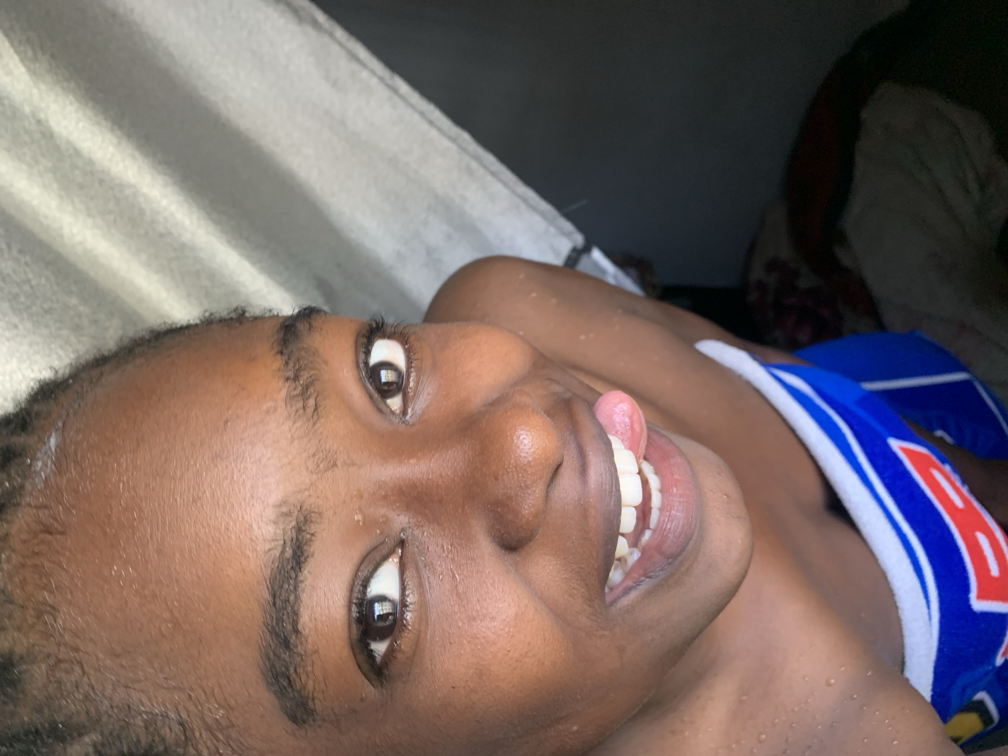 This media file is a close-up, low-angle portrait of a young person lying down. The subject has dark skin and is looking towards the camera with their tongue sticking out. They appear to be wearing a blue towel with white and red trim. There are water droplets visible on their skin, suggesting they may have recently bathed or been in a wet environment. The background is blurred and appears to be indoors, with a textured, light-colored surface on the left and darker, indistinct shapes on the right. The overall mood is playful and casual. There is no visible text in the image. The location context is Unguwar Saya, Nigeria.