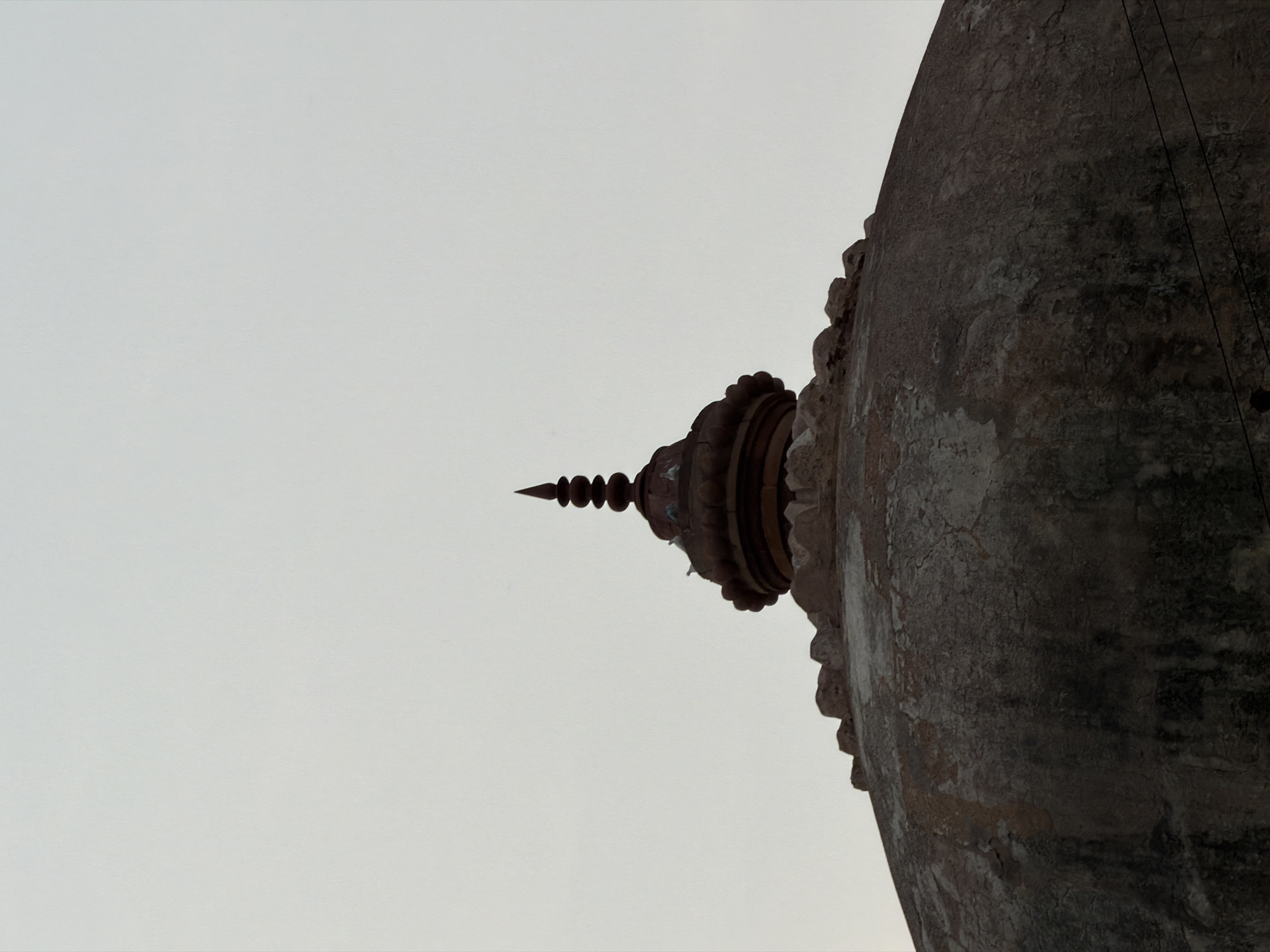 The image is a close-up, low-angle shot of the dome of a historical monument, likely a mosque or tomb, set against a pale, overcast sky. The dome is constructed from rough, textured stone, showing signs of age and weathering, with patches of darker discoloration and a cracked surface. At the apex of the dome, there is a tiered, spire-like finial made of dark, possibly metallic material, topped with a sharp point. Several birds, appearing to be pigeons, are perched on the decorative elements encircling the base of the finial. The upper edge of the dome features a crenellated or merlon-like border. The overall impression is one of ancient architecture viewed from below, emphasizing its scale and the passage of time. The image does not contain enough visual information to definitively place it in Delhi, India, or identify the specific monument.