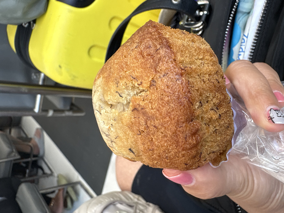 The image captures a close-up view of a banana muffin held by a human hand within an enclosed public setting in Pak Nam, Thailand.
**Subject:** A single, dome-shaped banana muffin with a golden-brown, textured crust and visible dark flecks, indicative of mashed banana or spices, occupies the central foreground. It is partially enclosed in a clear plastic bag and rests on a white, pleated paper liner.
**People & Action:** A human hand, with fingernails painted light pink, holds the muffin from below, gripping the plastic bag. The thumbnail displays a small, light-colored graphic, possibly a cartoon character. In the blurred background, the lower legs and feet of two other individuals are partially visible, one foot wearing a light grey slipper.
**Objects & Environment:** In the lower right background, a black bag or purse with silver-colored metallic rings and a zipper pull is partially visible. Inside this bag, a white and blue packet, possibly containing wet wipes, with partial text "Zaid..." is discernible. To the upper right, a bright yellow, contoured object with black straps, suggestive of a safety floatation device or a piece of luggage, is present. The background also reveals metal structural elements consistent with seating and a floor composed of dark grey or black material bisected by a distinct diagonal white or light grey stripe. This environment suggests the interior of a public conveyance or waiting area, such as a ferry, bus, or train, operating within Pak Nam, Thailand.
