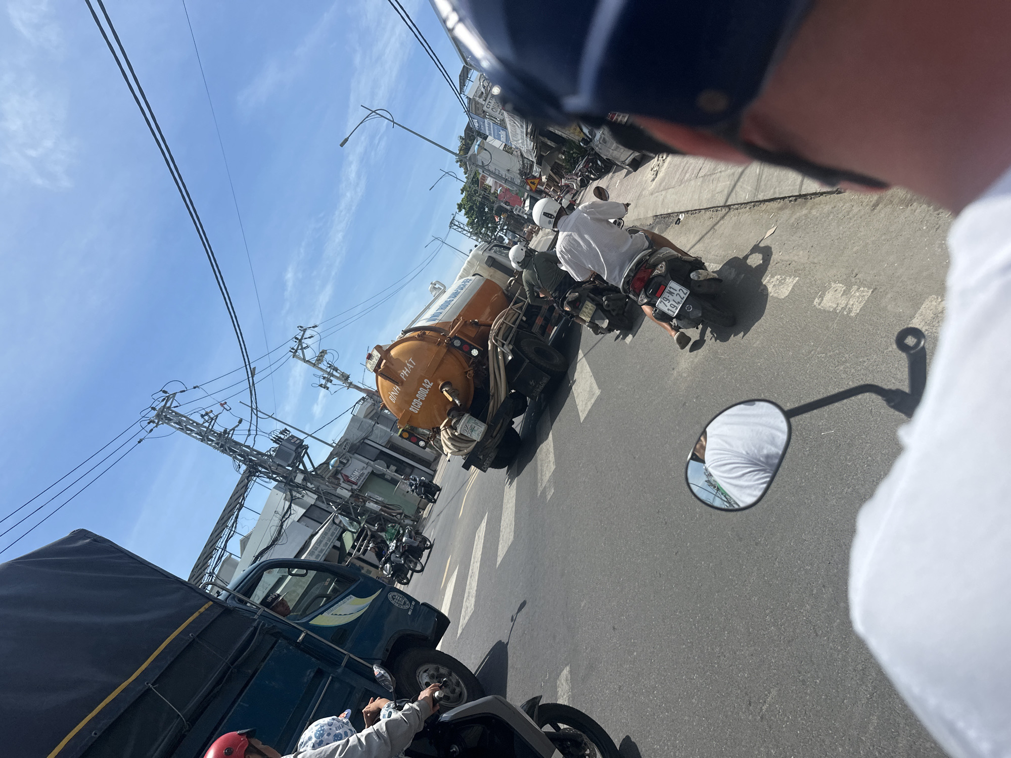 The image depicts a street scene in Bac Nha Trang Ward, Vietnam, under a clear blue sky with some scattered clouds. The foreground shows the back of a person's head wearing a dark helmet, partially obscuring the view. To the left, a white shirt is visible, suggesting the perspective of someone on a motorcycle.
In the midground, a dark blue truck with a black tarp is on the left, its front wheel visible. Next to it, an orange vacuum truck with "BINH PHAT" written on its side is moving forward. Several motorcycles are also present; one is dark colored with a license plate reading "79-N1 194.22," carrying two people wearing helmets. Another motorcycle is black and silver. A person in a white shirt and white helmet is riding a motorcycle.
The background reveals a street with buildings lining the right side, including signs for "HOME" and "CVH CTY TIEN." Power lines and poles are visible above the street, indicating urban infrastructure. The road is paved with asphalt and has white lane markings, including a pedestrian crossing. The overall scene suggests a busy street with typical urban traffic in Vietnam.