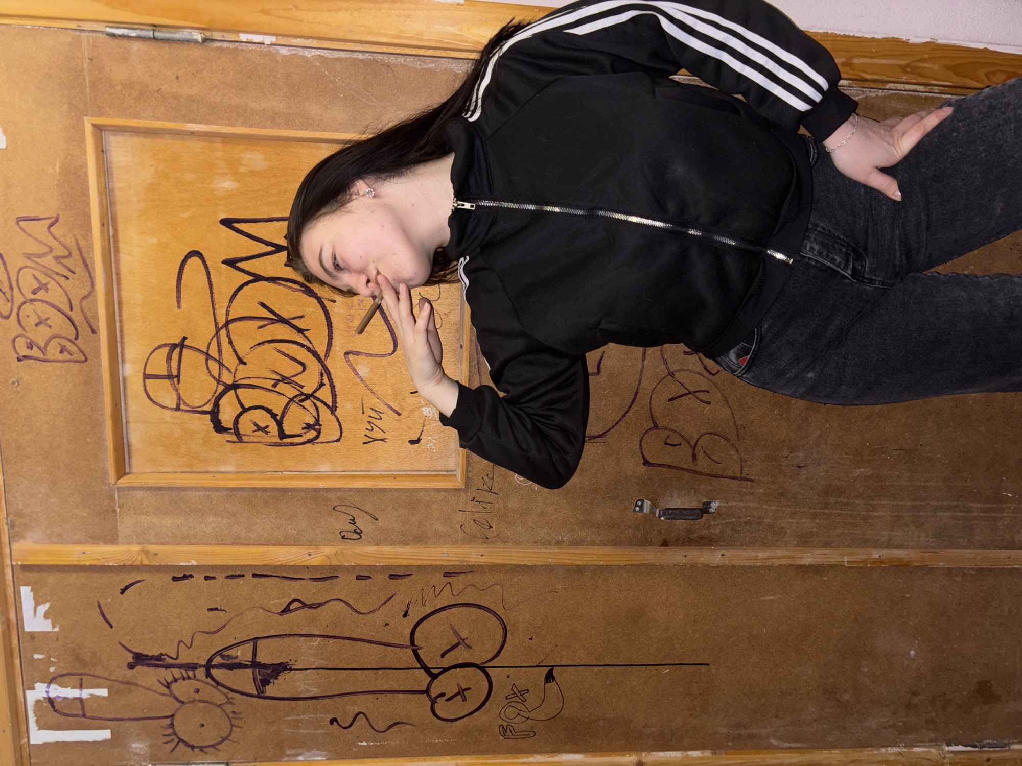 A young woman lies on a door covered in graffiti. She is wearing a black tracksuit with white stripes on the sleeves and dark gray jeans. She has long, dark hair. She is holding a rolled cigarette to her lips. The door has various graffiti markings, including drawings of genitalia. The city of Perm, Russia is not apparent in the image.