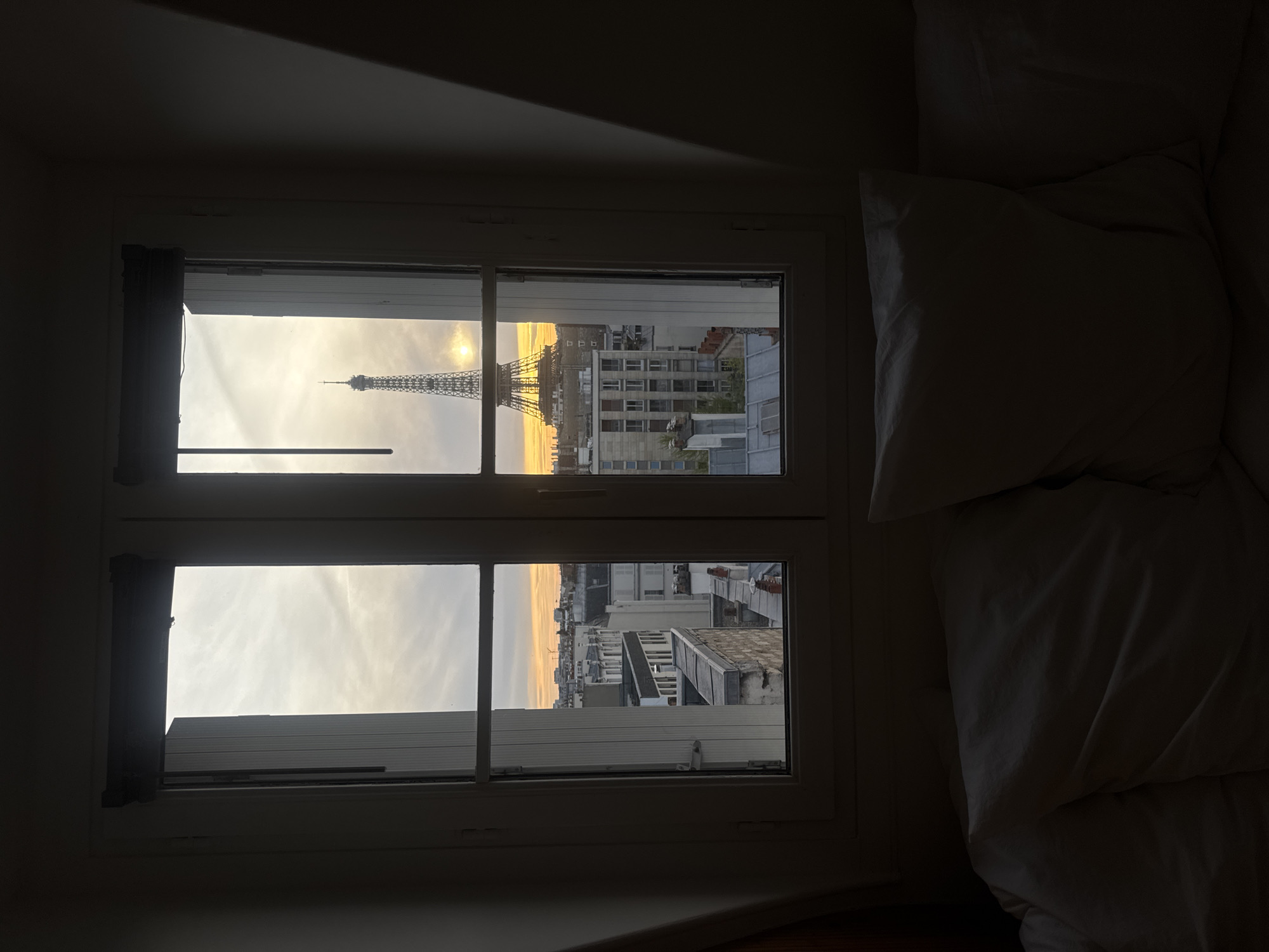A view of the Eiffel Tower in Paris, France, seen through an open window from an interior bedroom. The sky indicates it is either sunset or sunrise. A bed with white pillows and bedding is situated to the right of the window. The bedroom is dimly lit. Buildings can be seen in the distance to the right of the Eiffel Tower.