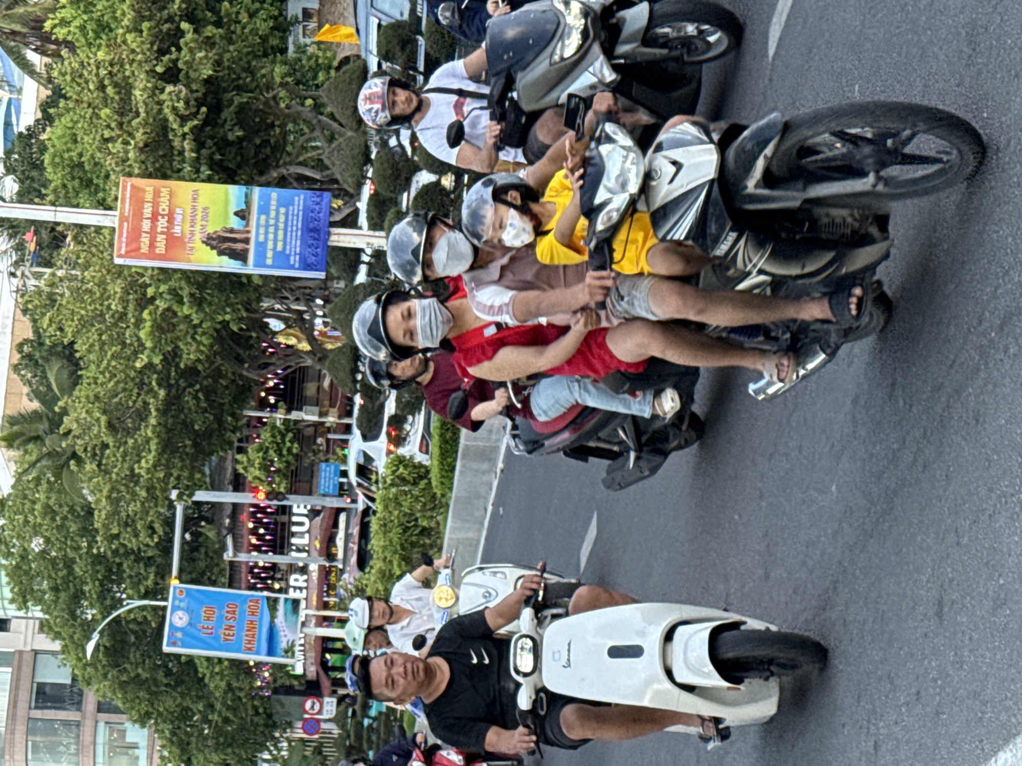 This is a vertical, eye-level, outdoor photograph taken in Nha Trang, Vietnam. The image captures a busy street scene with multiple motorbikes and scooters carrying passengers. The overall impression is one of vibrant street life and local activity.
The primary subjects are the people riding the motorbikes, most of whom are wearing helmets and some are wearing face masks. The people are of various ages, and the groups suggest families or friends traveling together. They are depicted in motion, suggesting they are either commuting or participating in a local event.
Objects in the scene include several motorbikes and scooters, their designs varying from sleek modern scooters to more rugged-looking motorbikes. Street signs are also visible, including one in Vietnamese that appears to be advertising an event related to Cham culture and the Khanh Hoa province, possibly a festival titled "NGÀY HỘI VĂN HOA DÂN TỘC CHĂM LẦN THỨ VI TẠI TỈNH KHÁNH HOA NAM 2026". Another blue sign reads "LỄ HỘI YẾN SAO KHÁNH HOA". Green foliage from trees lines the street, and buildings can be seen in the background, indicating an urban setting.
The time of day appears to be late afternoon or early evening, as suggested by the lighting and the warm hues in the sky, although this is not explicitly clear. The weather seems pleasant, as people are not wearing heavy clothing.
The overall mood is lively and dynamic, reflecting the hustle and bustle of Vietnamese street traffic. The presence of multiple people on each vehicle highlights a common mode of transportation and social interaction in the region. The vibrant colors of the signs and clothing add to the visual energy of the scene.