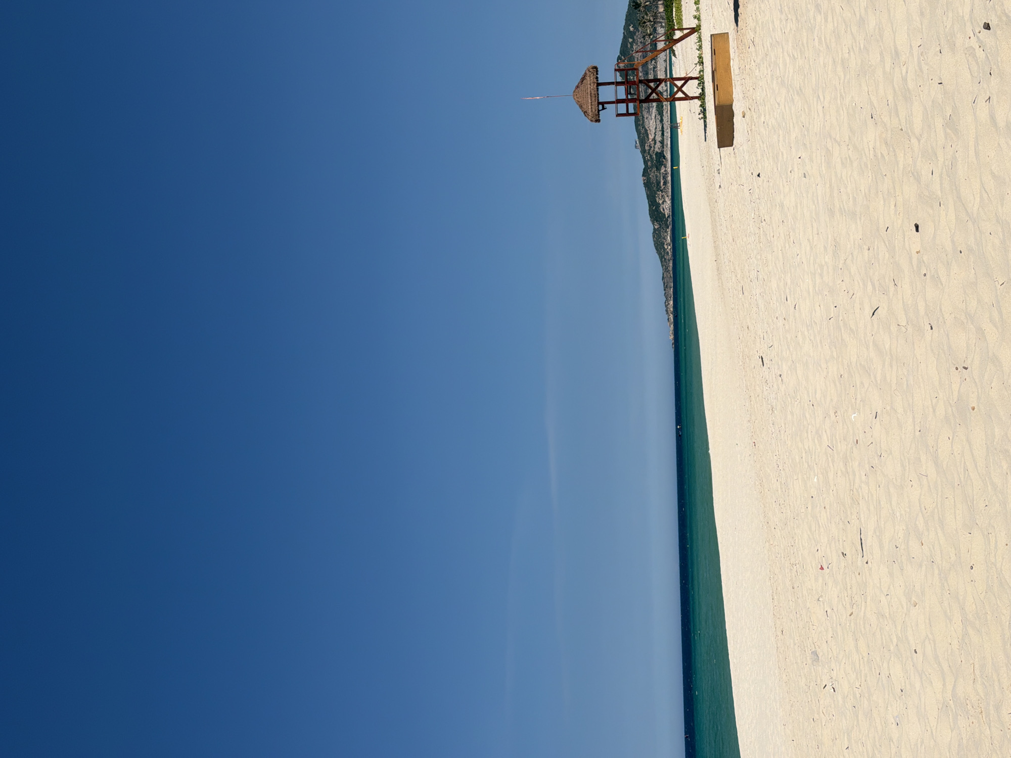 A wide, sandy beach stretches from the foreground to meet a calm, turquoise ocean under a clear, bright blue sky. A lifeguard tower with a thatched roof stands on the right side of the beach, its wooden structure visible against the distant green, hilly landscape. A narrow strip of darker blue water borders the turquoise shallows, and faint landmasses are visible on the horizon. The sand is a light, pale yellow, with some small debris scattered across its surface. The overall scene depicts a tranquil, tropical beach environment.