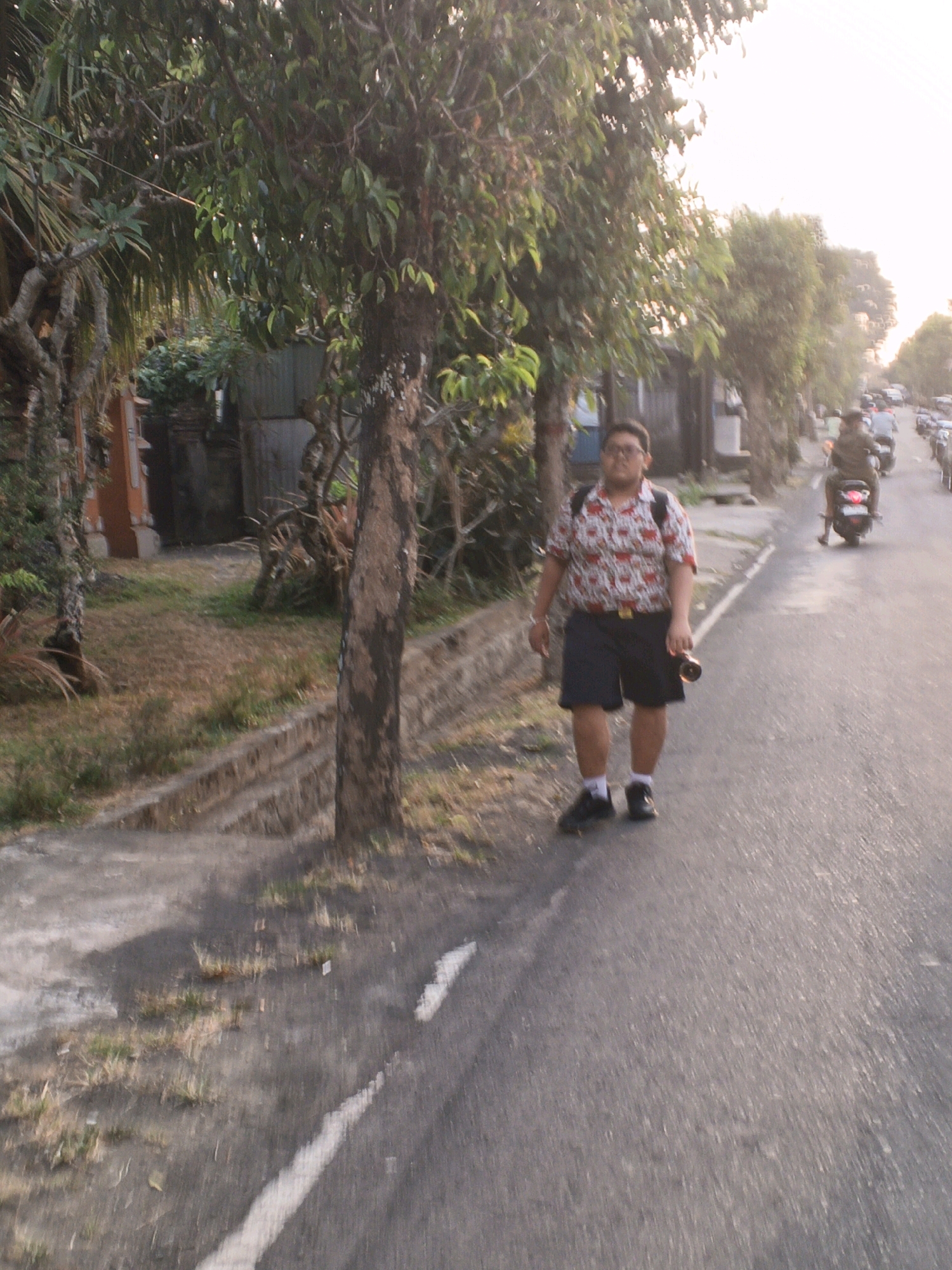 A young person wearing glasses, a short-sleeved patterned shirt, and dark shorts walks on the side of a paved road in Sukawati, Indonesia. They have a backpack on their shoulders and are holding a dark object in their right hand. The person is walking towards the viewer, who is positioned on the left side of the road. The road is lined with trees on the left, with buildings and more vegetation visible behind them. On the right side of the road, other people are seen on motorcycles, and a line of parked vehicles is further down the road. The sky is bright and appears to be either early morning or late afternoon. The ground on the left side of the road consists of dry grass and a concrete drainage ditch. The overall lighting suggests a bright, sunny day.