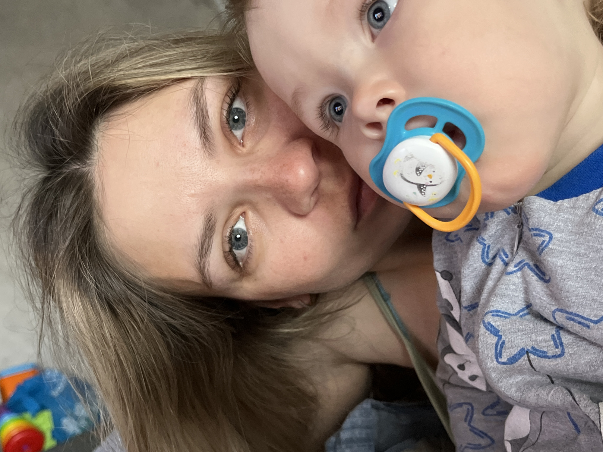A close-up selfie shows a fair-skinned woman and a baby, presumably in a home setting. The woman has light brown, shoulder-length hair with blonde highlights and blue eyes. The baby is fair-skinned with blue eyes and has a blue and orange pacifier in its mouth. The baby is wearing a gray patterned pajama shirt. The background is out of focus. There is no indication of the location being the city of Chaikovsky, Russia.