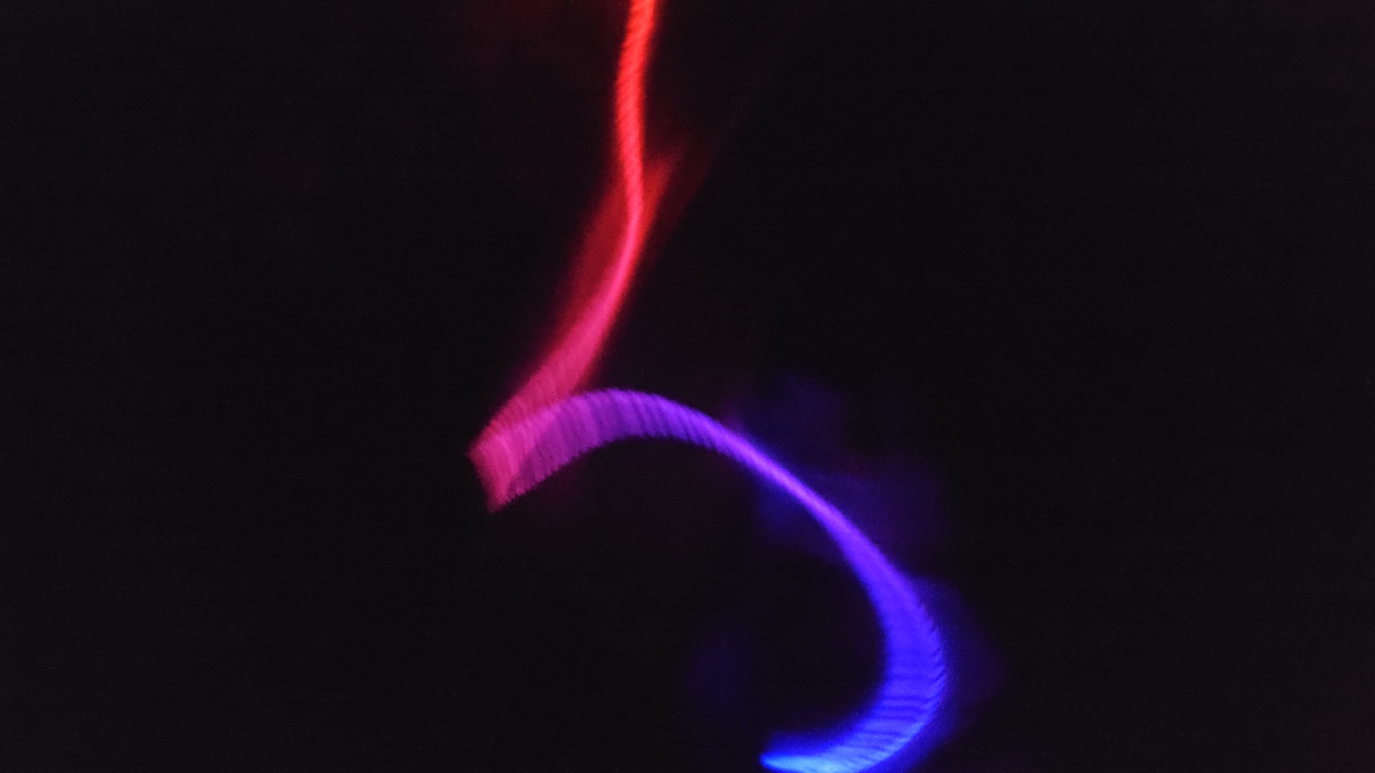 The image is a long-exposure photograph showing blurred streaks of red and blue light against a dark background. The red light appears as a vertical line at the top, curving slightly to the left. Below it, the blue light forms a wider, curved shape, transitioning through shades of purple where the colors blend. The background is black with faint speckles of light. There is no discernible information in the image to indicate its location as Cam Đức, Vietnam, or any other specific place. There are no people or distinct objects visible. The image appears to be an abstract representation of light movement.