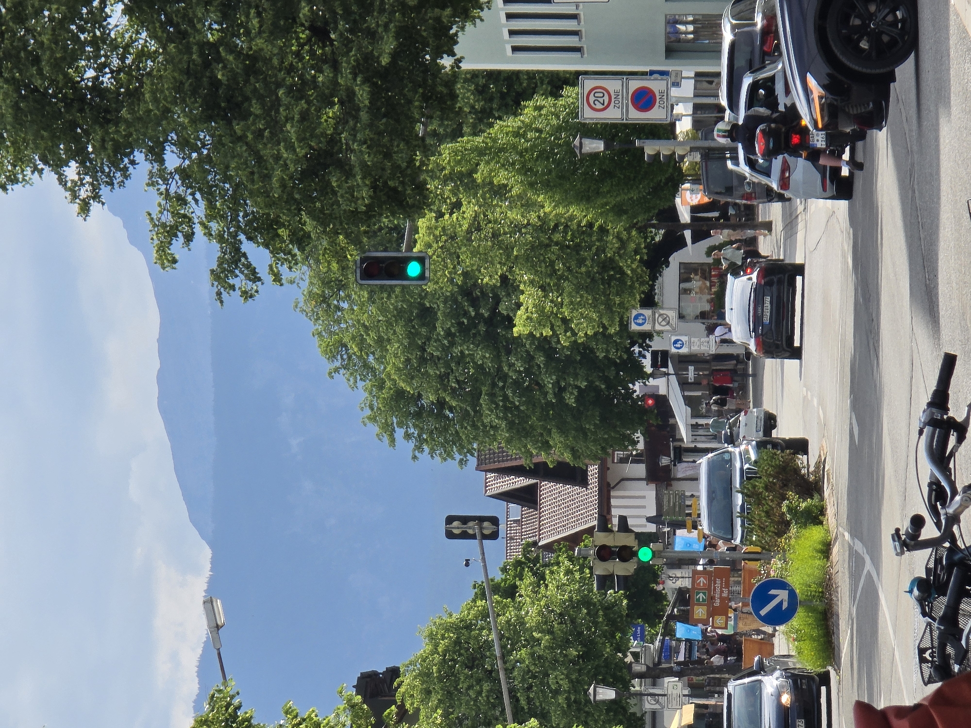 The image depicts a street scene in Garmisch-Partenkirchen, Germany, on a clear, sunny day. In the foreground, the handlebars and front wheel of a bicycle are visible. The middle ground shows a street with several cars, including a black Audi, a white SUV, and a motorcycle with a rider. Traffic lights are visible, with one showing a green light. Buildings with pitched roofs line the street, and trees with lush green foliage are abundant. In the background, a mountain range with a hazy blue hue is visible under a partly cloudy sky. Several road signs are present, including a "20 ZONE" sign and a sign with a pedestrian symbol. People are seen walking on the sidewalk and near the buildings. The overall impression is of a busy town center in a scenic mountainous region.