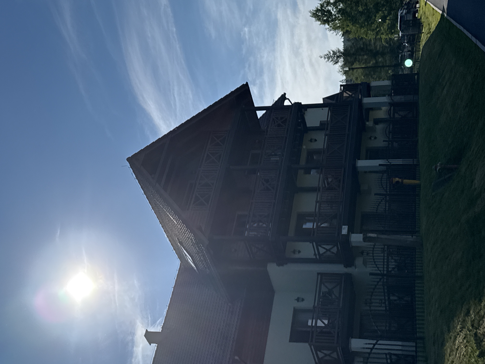 The image depicts a multi-story wooden and stucco building with multiple balconies, situated in Szklarska Poręba, Poland. The sun is prominently visible in the upper left corner of the frame, casting a lens flare and illuminating the clear blue sky with wispy white clouds. The building features dark wooden balconies with intricate criss-cross detailing, and a dark-tiled roof. A black metal fence runs along the front of the property, separating it from a grassy slope. In the background, dense evergreen trees line a hillside, suggesting a mountainous or forested area. To the right, a paved path runs alongside the property, with parked vehicles visible beyond the fence, indicating a road or parking area. The overall scene suggests a serene, sunlit day in a natural, possibly resort-like setting.