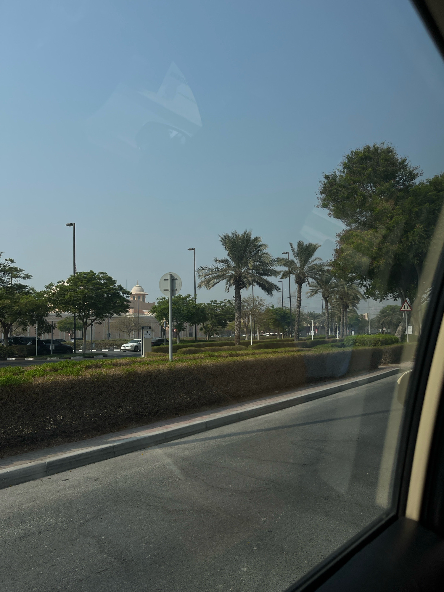 The image depicts a view from inside a vehicle in Dubai, United Arab Emirates. The foreground shows the asphalt road and a concrete curb, with a dense green hedge lining the roadside. Beyond the hedge, a paved area with lampposts and several parked cars is visible. In the middle ground, a grand building with a dome and arches stands prominently, surrounded by trees and palm trees. The sky is clear and blue, with reflections from the vehicle's windows visible on the surface of the image. A traffic sign with a U-turn symbol is also present on the right side of the road. The overall scene suggests a modern, well-maintained urban environment in Dubai, possibly a public space or a government building.