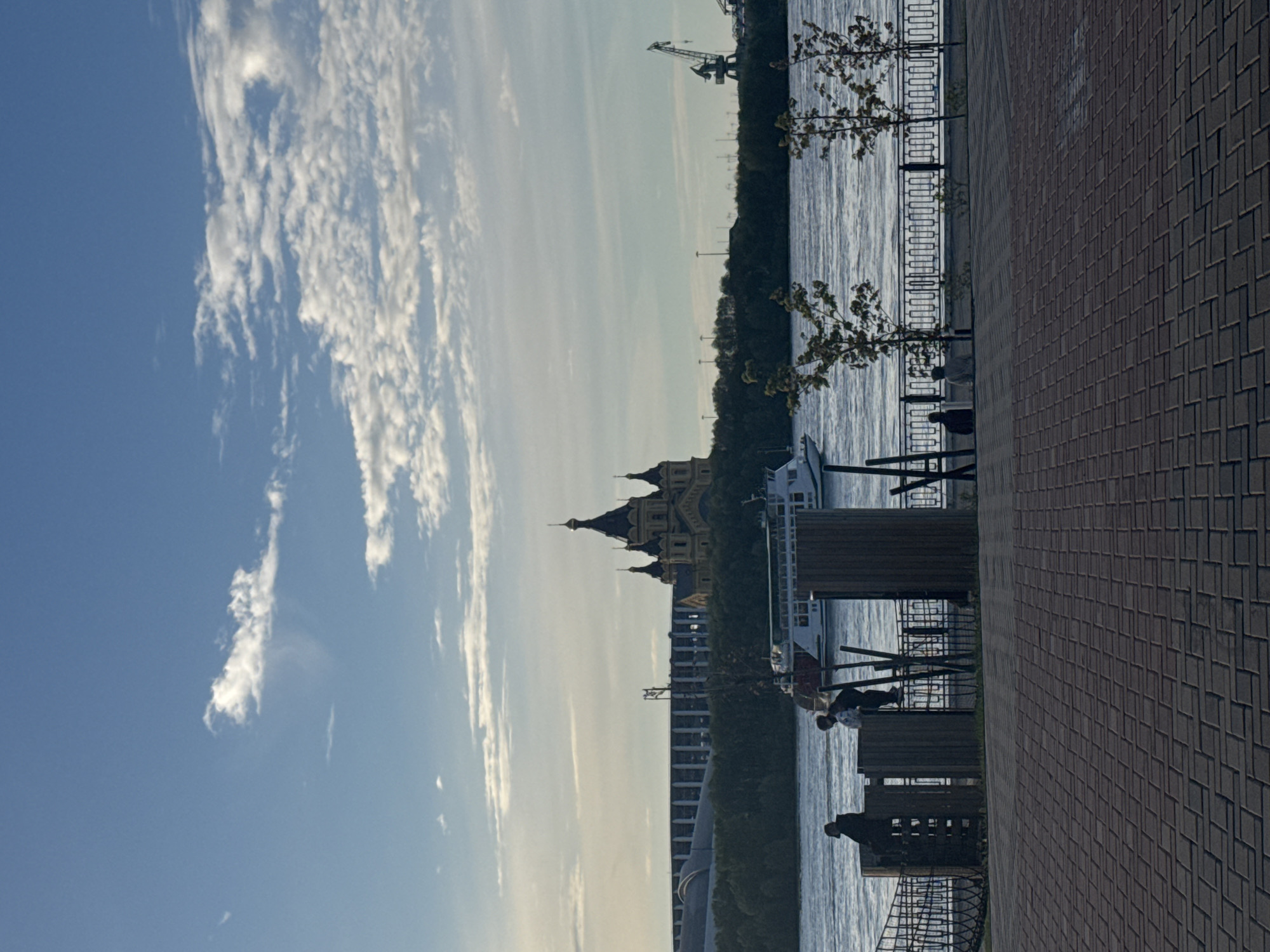 The image shows a view of Nizhny Novgorod, Russia. A brick-paved walkway lines the right side of the image. A black, wrought-iron fence separates the walkway from a body of water, likely the Volga River. On the water, a white boat is visible. Across the river, a forest is visible, partially obscuring a building with a dark roof and multiple ornate domes. To the left of the building, a series of high-rise apartment buildings is seen. A crane is visible in the distance on the right side of the image. Above, a blue sky with long, thin clouds dominates the left side of the image. Two people appear to be seated on benches along the walkway.
