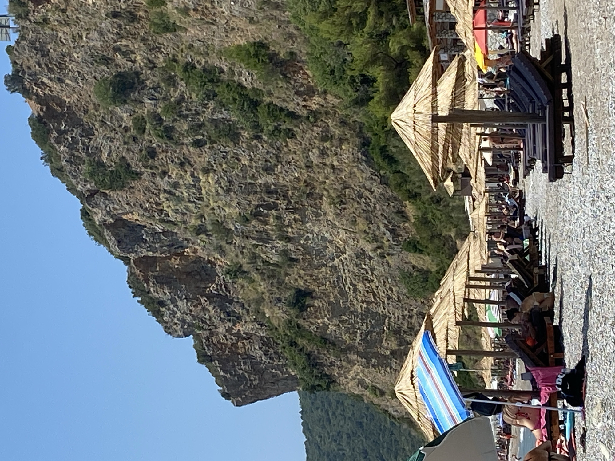 The image depicts a pebble beach in Prijevor, Montenegro, on a clear, sunny day. A large, rocky cliff face dominates the background, with sparse vegetation growing on its slopes. In the foreground, rows of lounge chairs and thatched umbrellas are arranged on the beach, suggesting a tourist resort area. Several people are visible relaxing on lounge chairs, some partially obscured by umbrellas. The sea is visible in the distance, with gentle waves. The overall impression is one of a tranquil beach scene.