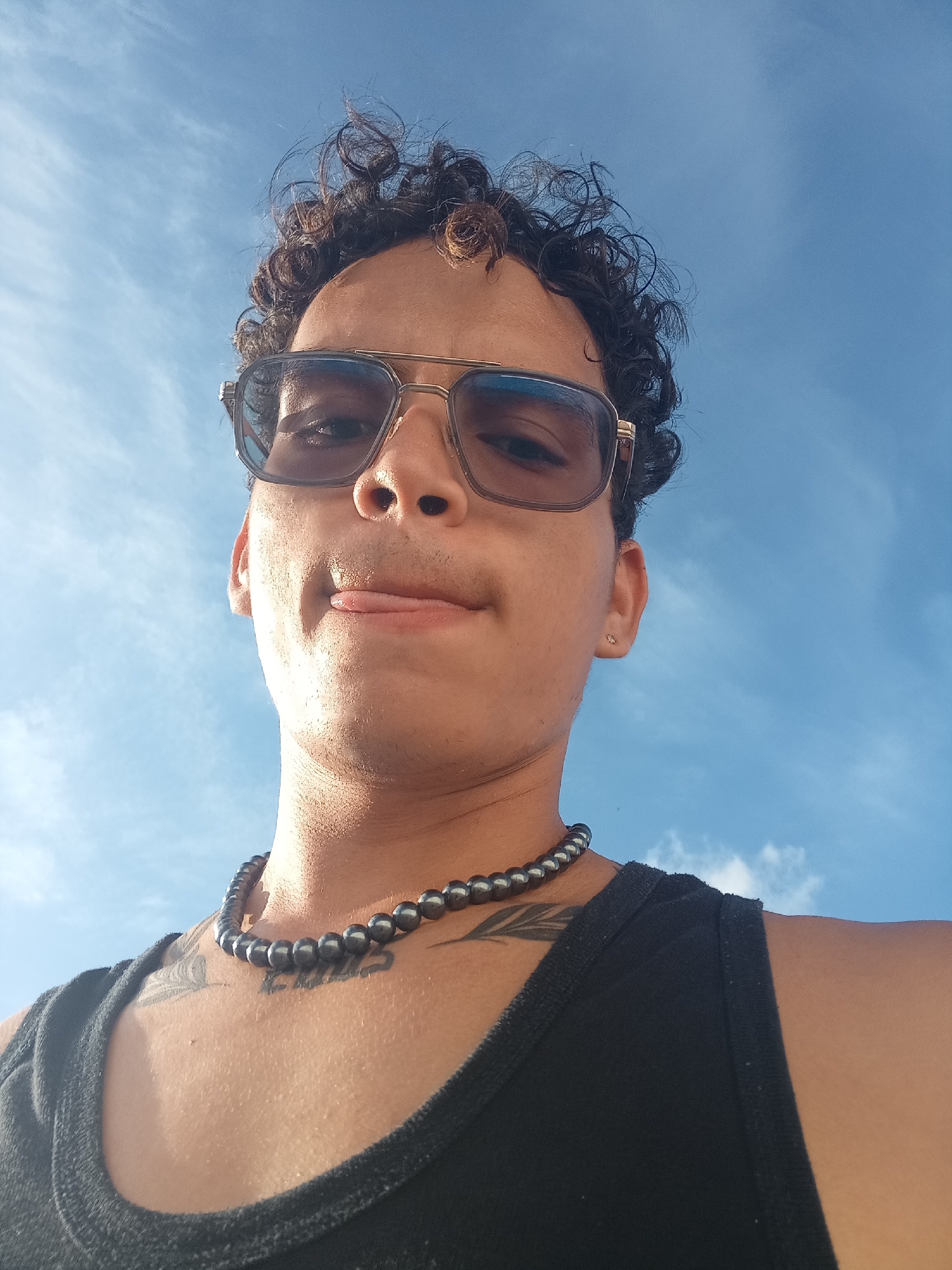 A low-angle, outdoor, medium shot captures a person's face and upper torso against a bright blue sky with wispy white clouds. The subject, viewed from below, wears large, aviator-style sunglasses with a gold frame and tinted lenses. Their curly dark hair is windswept. The person has their tongue sticking out slightly between their lips. Around their neck, they wear a dark, beaded necklace. A black tank top is visible on their torso, with tattoos on their chest and shoulder. The lighting suggests it is daytime, possibly sunny. The image's perspective, looking upwards at the subject against the sky, is reminiscent of the viewpoints often found in coastal areas or open public spaces. Given the reference to Juan Griego, Venezuela, the scene could be a beach or a seaside promenade.