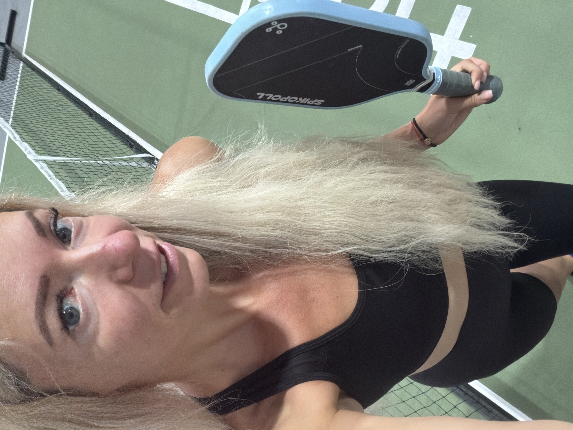 A blonde woman in athletic wear, holding a light blue pickleball paddle, is positioned on an indoor pickleball court with a green surface and a black net visible in the background. The woman is looking towards the camera with a slight smile. The image is a close-up, focusing on the woman's upper body and the paddle she is holding. The scene is indoors, and there is no indication of the city of Da Lat, Vietnam.