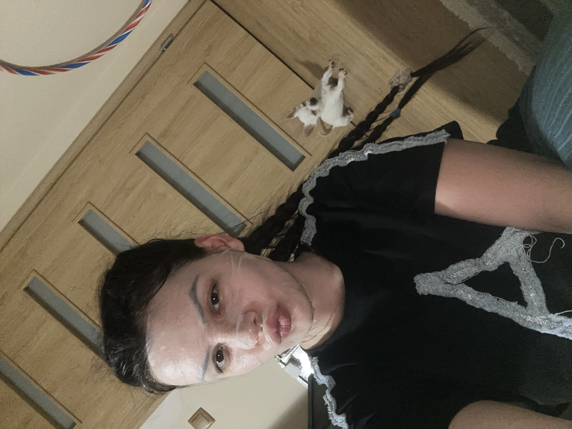 A person with a facial mask lies sideways in a room. The person has dark hair braided into two pigtails. A small white and black dog lies on the wooden surface behind the person. The person wears a black shirt. A red, white, and blue hoop lies in the upper left corner of the image. The location is Chubra, Bulgaria.