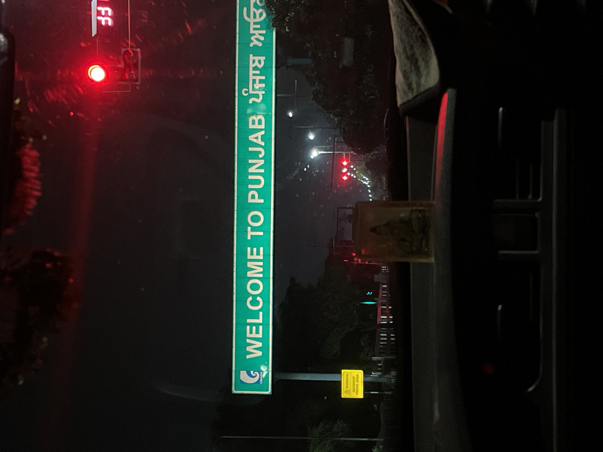 The image captures a nighttime view from inside a vehicle, looking forward. Dominating the scene is a large green overhead sign with white lettering that reads "WELCOME TO PUNJAB" in English, followed by text in Punjabi script. A logo for "GMADA" is visible on the left side of the sign. To the left of the sign, a yellow warning sign with a black border and symbols is attached to a pole. The environment is dark, indicating nighttime, with streetlights and traffic signals visible in the distance, including a red traffic light. Trees and foliage line the roadside. The interior of the vehicle is visible in the foreground, including a dashboard and a small religious figurine placed on it. The overall scene suggests the vehicle is at or near a border crossing into Punjab, possibly on a highway or major road leading into or out of Mohali, India.