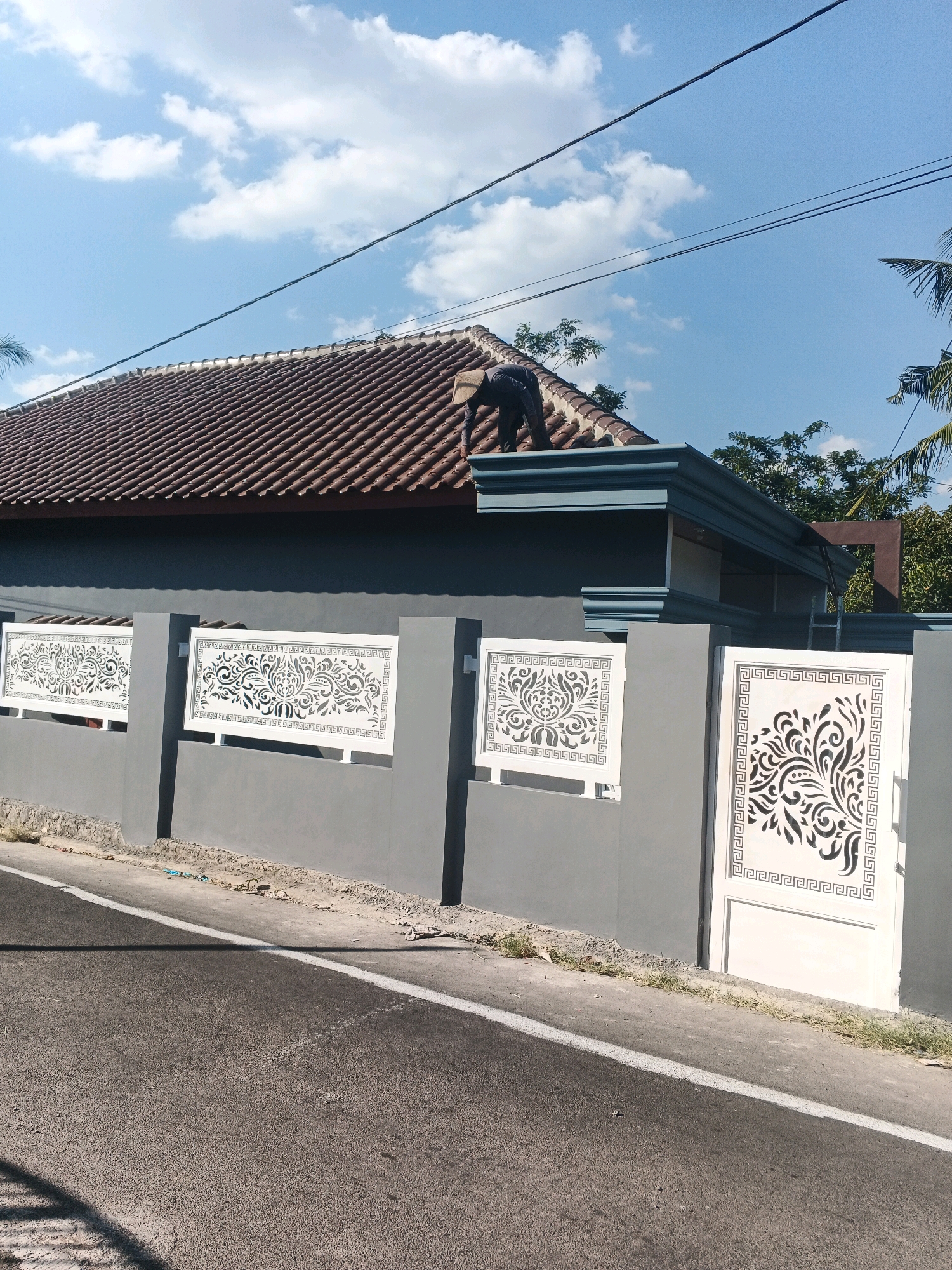 In Ungaran, Indonesia, a person in a light-colored hat and dark clothing is on the roof of a house, appearing to be performing maintenance or repairs. The house has a brown tile roof and a gray wall with decorative white panels. A white gate with an intricate black pattern is also visible. The scene is set against a bright blue sky with scattered white clouds, and trees are visible in the background. A paved road runs in front of the property.