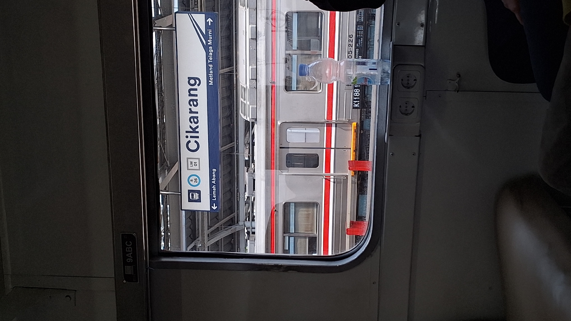 The image, taken from inside a train, shows a view through a train window of another train at a station. A blue sign indicates the "Cikarang" station, with directional information for "Lemah Abang" and "Metland Telaga Murni". A bottle of water is placed on the windowsill. Power outlets are visible on the wall next to the window. The serial number "K11881" is visible on the adjacent train. The train windows are reflecting.