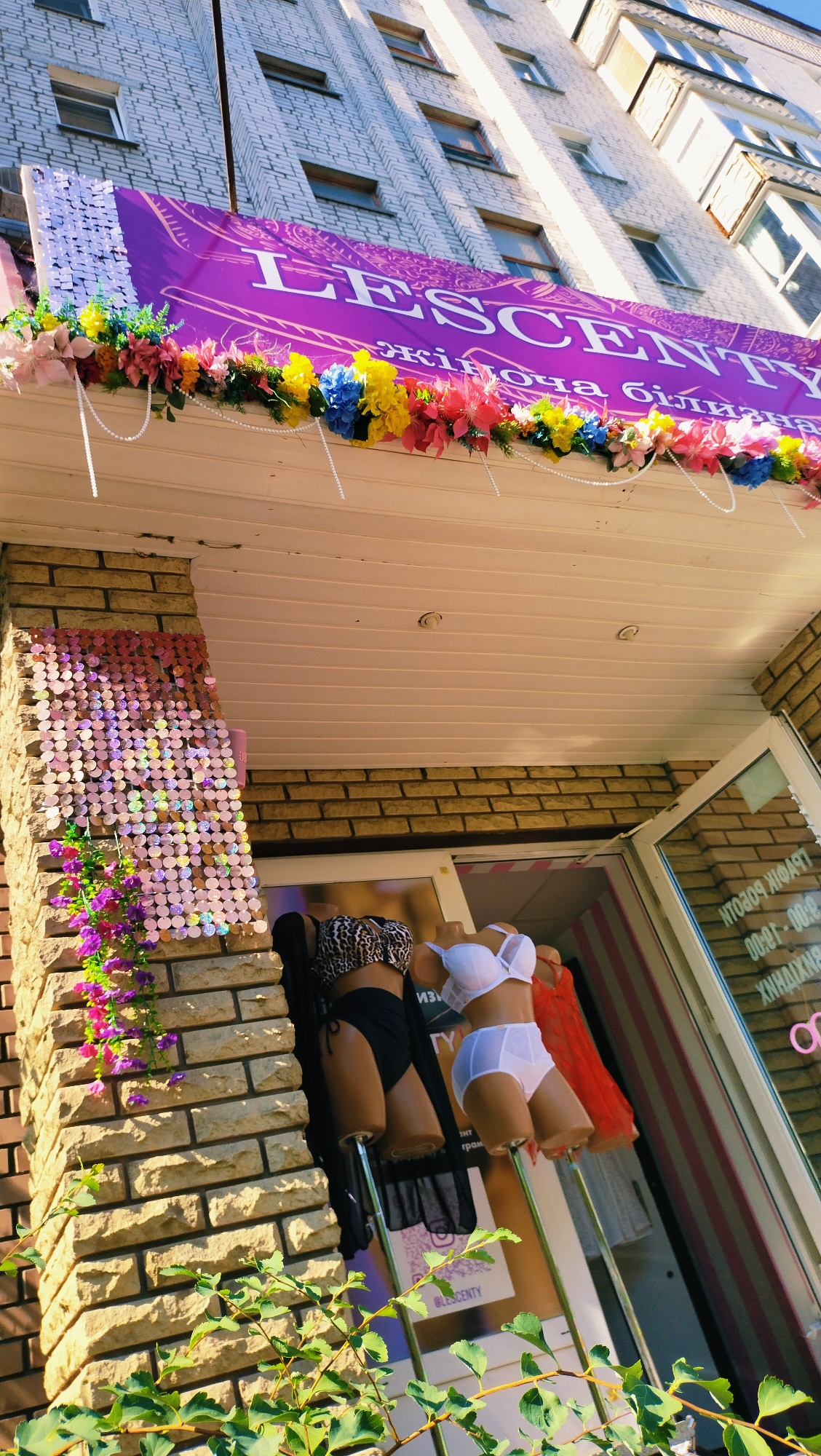 The image shows the exterior of a lingerie shop named "BLESCENTY" located in the city of Horishni Plavni, Ukraine. The shop has a purple banner with white lettering and is decorated with colorful artificial flowers and pearl garlands. In front of the store, mannequins are displayed wearing lingerie: one mannequin wears a leopard print bikini and a black sheer robe, and another wears a white bra and underwear set. The store entrance is visible with a glass door and a sign with Cyrillic text. The building is made of brick and has a white ceiling above the entrance. Green foliage is in the foreground, partially obscuring the view.