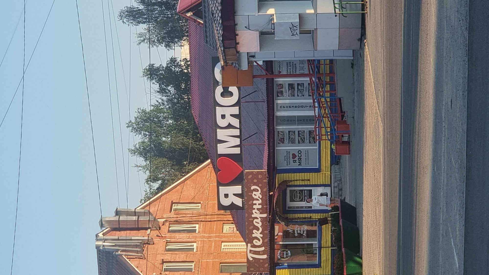 This is a photograph taken in the city of Megion, Russia, of a building with storefronts. Dominating the scene is a large, illuminated sign that reads "Я ♥ МЯСО" (Ya Lyublyu Myaso), which translates to "I Love Meat." Below this sign is a storefront with a yellow facade and a brown roof, advertising "Pekarna" (Bakery). Another storefront to the right has a sign that also includes "Я ♥ МЯСО" and phone numbers. A person in a white t-shirt and shorts stands near the entrance of the bakery, holding a bag. The environment appears to be a commercial area with trees visible in the background and power lines overhead. The time of day seems to be late afternoon or early evening, as indicated by the lighting.