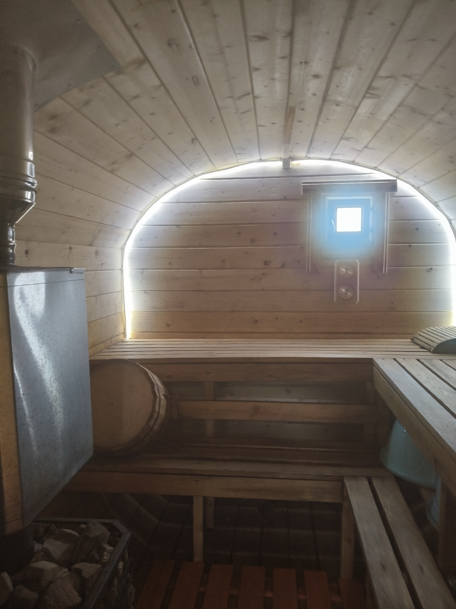 The image depicts the interior of a sauna in Chaikovsky, Russia. The sauna has wooden walls and ceiling with an arched top, and a small window. Inside is a stove with stones, a water bucket, and wooden benches. There are no people present in the sauna.