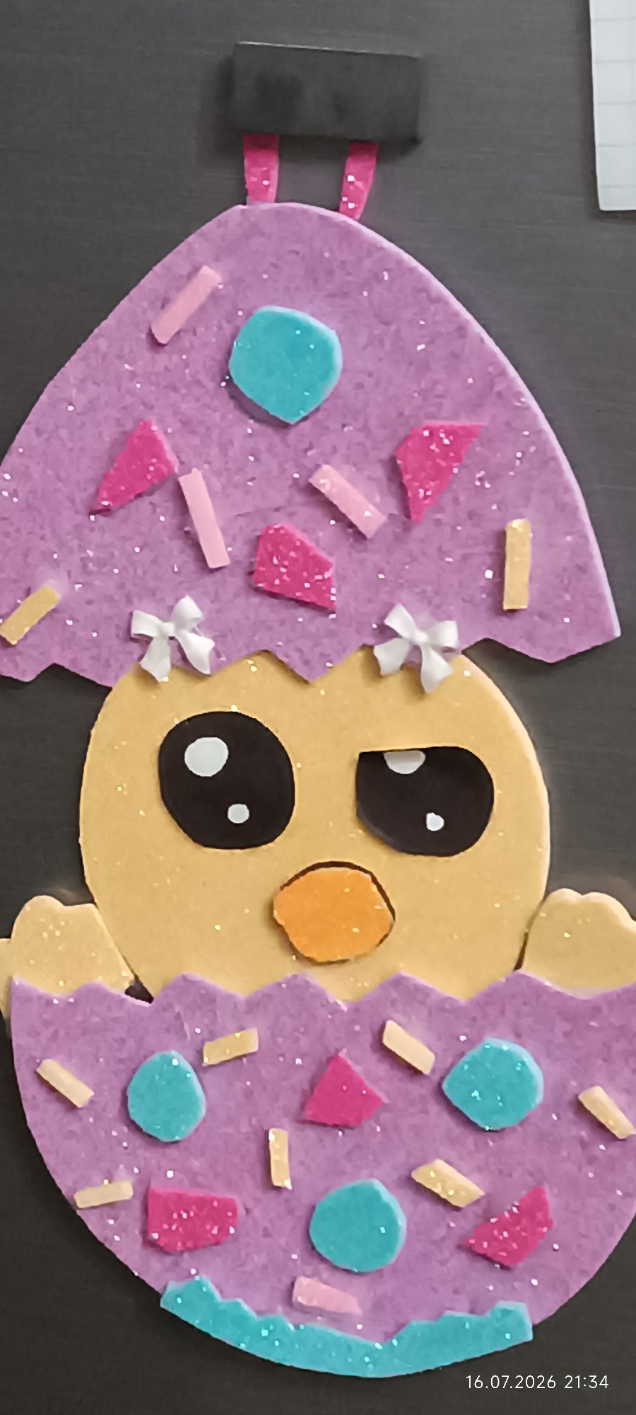 This is a close-up, top-down shot of a handmade craft project, likely for Easter. The primary subject is a cartoonish yellow chick emerging from a cracked purple egg. The chick has large black oval eyes with white dots for pupils, and a small orange beak. It is adorned with two small white bows on the upper part of the eggshell.
The eggshell, made of purple glitter foam, is decorated with various small, colorful shapes including light blue circles, pink triangles, and light pink and yellow rectangular pieces, resembling sprinkles or confetti. The bottom portion of the eggshell is also purple and similarly decorated. The craft is placed on a dark, textured surface, possibly a tabletop or desk.
A timestamp in the bottom right corner reads "16.07.2026 21:34", suggesting the image was taken on July 16, 2026, at 9:34 PM.
There is no discernible connection to the city of Odesa, Ukraine, in this image.