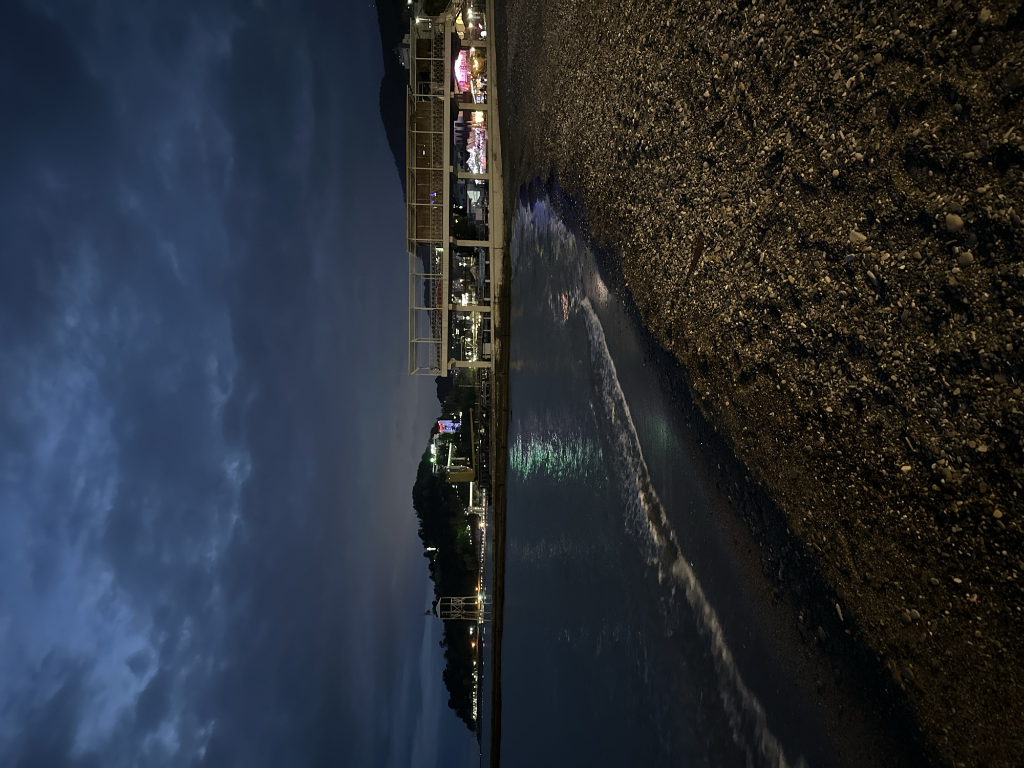 This night scene captures a lively coastal area in Sochi, Russia. The image, oriented as if looking along a coastline, depicts a dark, cloudy sky above a landscape featuring illuminated buildings, a body of water, and a pebbly beach.
In the upper left portion, a dark blue-grey sky is visible, heavy with clouds, suggesting an overcast night. Below this, distant mountains or hills are silhouetted against the faint light, forming a natural backdrop to the urban stretch.
The central part of the image is dominated by a promenade or boardwalk lined with various buildings, brightly lit with artificial lights. Some structures feature open-air designs with white trellised frameworks, possibly indicating restaurants or cafes. Patches of vibrant pink, yellow, and white lights from these establishments are prominent, reflecting off the water. Further along the coastline, more clusters of lights suggest additional buildings and activity, indicating a developed area.
To the right, a dark, pebbly beach stretches into the foreground, composed of countless small, dark stones. Gentle waves break on the shore, their white foam contrasting with the dark water. The water itself, likely the Black Sea, reflects the colorful lights from the shore, creating shimmering streaks of green, blue, and purple across its surface. No specific human activity is discernible, but the illuminated structures suggest a bustling evening atmosphere typical of a resort town. The overall impression is one of a calm yet vibrant night by the sea. No visible text is legible.