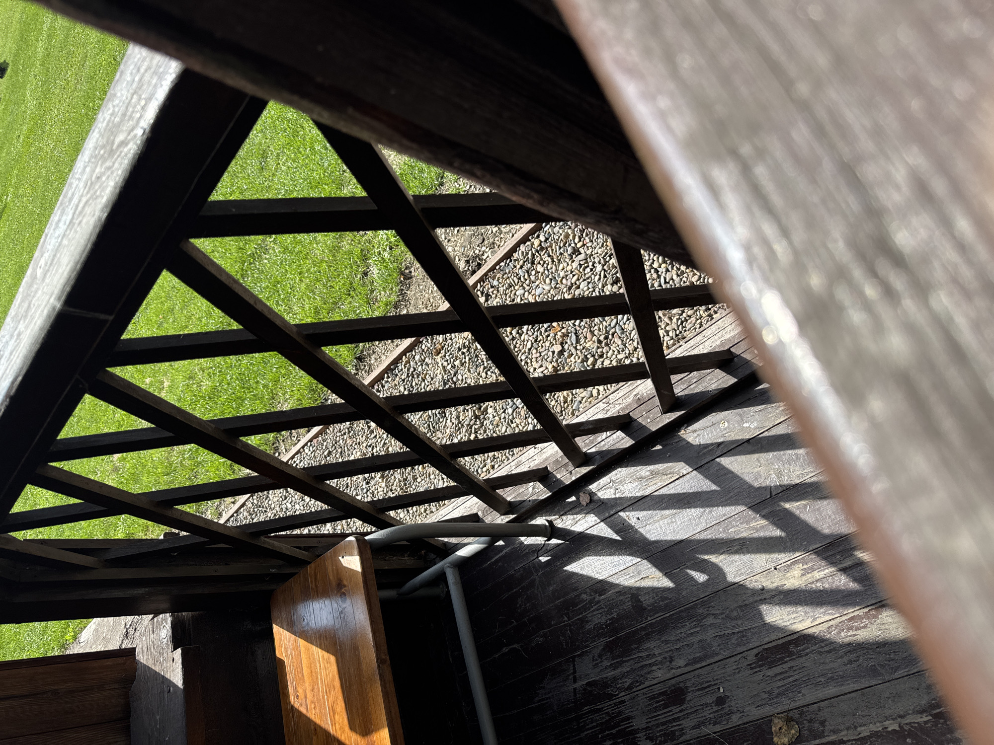 The image depicts a wooden porch or balcony with a railing and a wooden bench. The porch floorboards are weathered and dark. Sunlight casts a geometric shadow pattern on the floor from the railing's diamond-shaped lattice. Beyond the railing, there is a gravel area and a neatly manicured green lawn, suggesting an outdoor setting. The overall impression is one of a tranquil, possibly rural, or suburban environment. The specific location in Ust-Mana, Russia, is not visually indicated by any landmarks or text within the image itself.