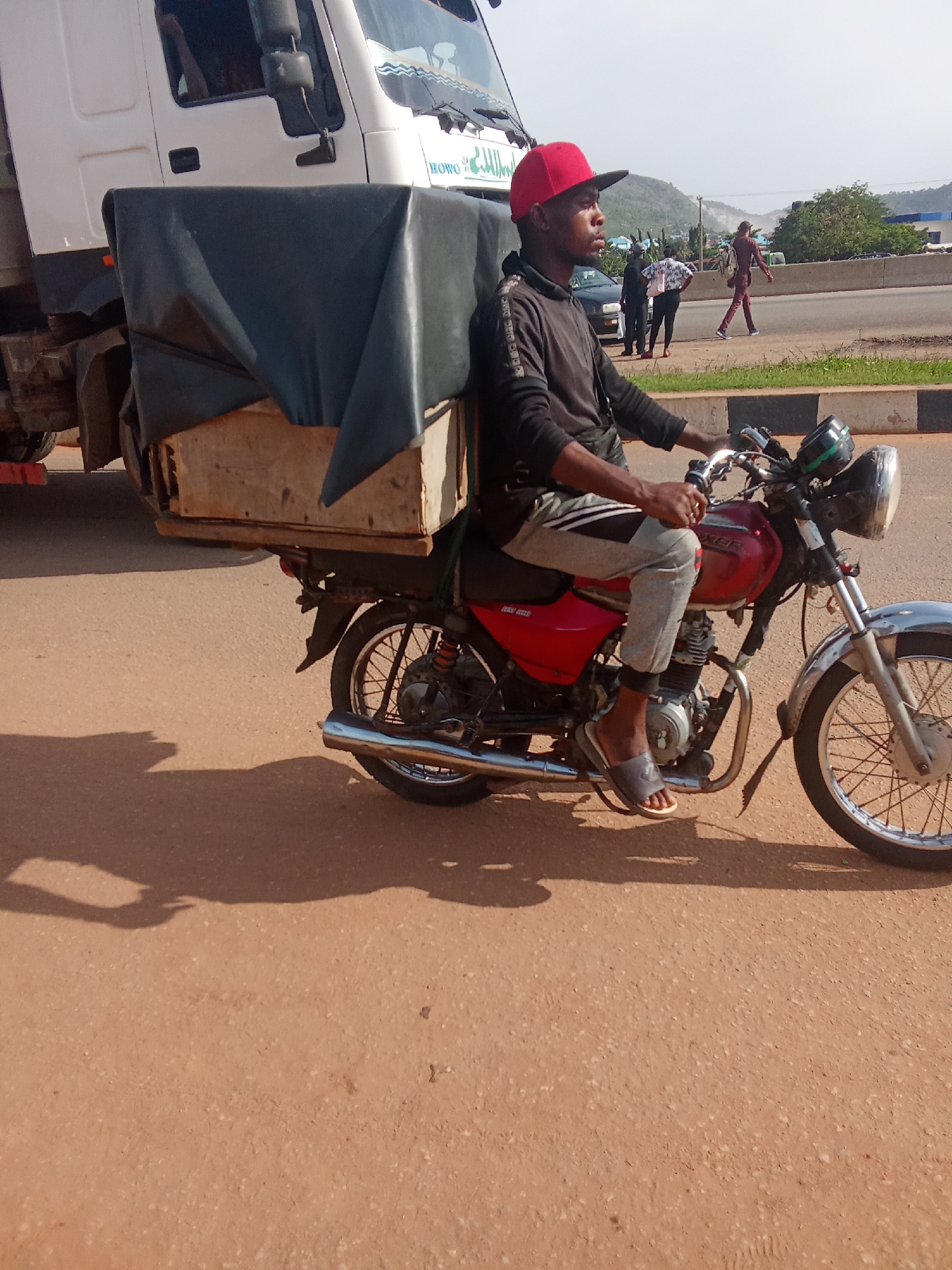 A man wearing a red baseball cap, a dark hoodie, and grey sweatpants rides a red motorcycle. He is looking to his left. The motorcycle is a Bajaj Boxer 150. The man is wearing grey flip-flops. In the background, there is a white truck with a black tarp over a wooden box. The truck appears to be parked. Other people are visible walking on the side of a road behind the motorcycle. The scene appears to be in a city with a mountainous background. The road is paved with a reddish-brown asphalt. The sky is clear and bright.