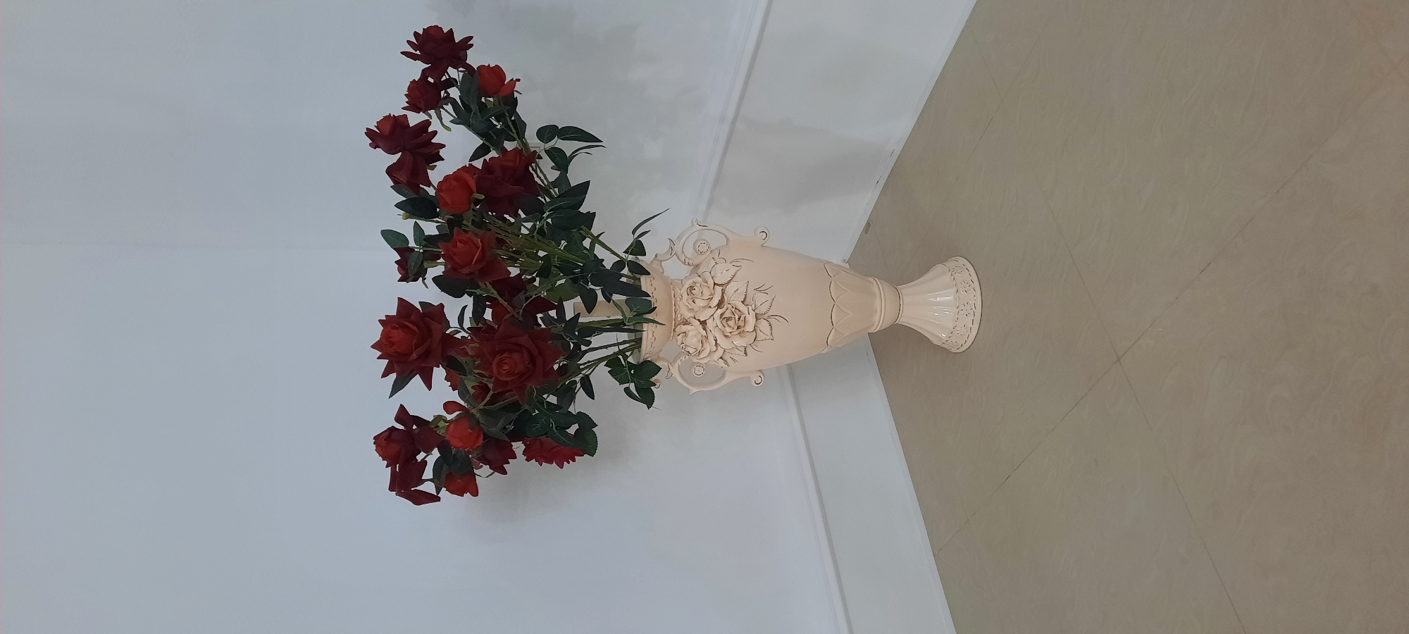 A bouquet of artificial red roses is displayed in an ornate, cream-colored ceramic vase. The vase has a wide opening and is decorated with raised floral patterns and scrolling handles. The roses are a deep red, with some appearing more open and others tightly budded, interspersed with green artificial leaves. The arrangement is positioned in a room with a white wall and a light-colored tiled floor. The tiles have a subtle pattern resembling wood grain and are laid out in a grid with visible grout lines. The scene appears to be indoors, with artificial lighting. There is no indication of the time of day or weather. The location context is Gudermes, Russia, but no specific cues within the image support this. There is no text visible in the image. The overall impression is one of a decorative still life, possibly in a home or a commercial space like a hotel lobby or shop. The lighting is soft and even, suggesting an interior environment.