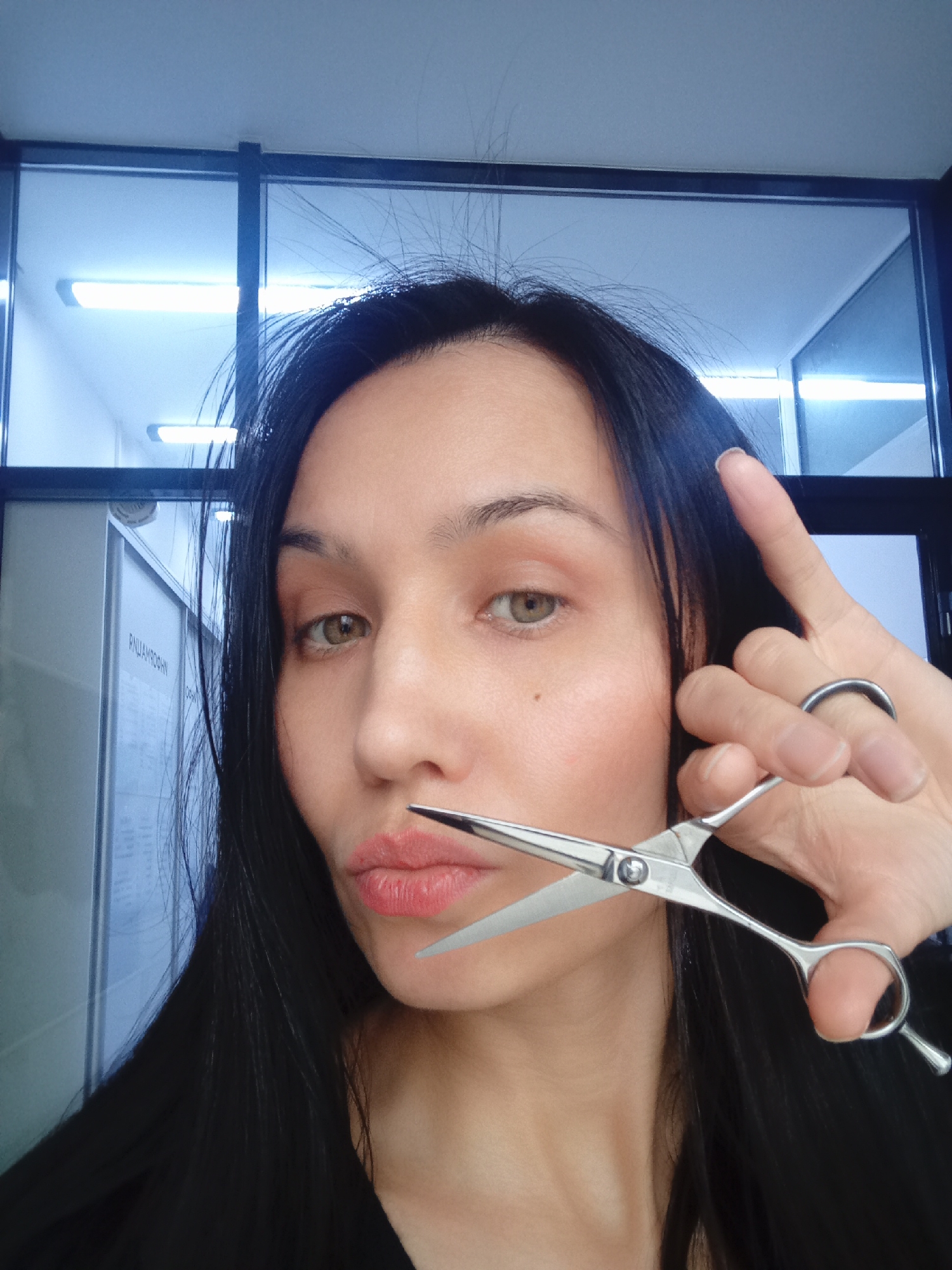 A woman is holding a pair of open scissors near her mouth in a selfie-style photograph. Her hair is long, dark, and straight. She has fair skin, light eyes, and is wearing lipstick. The background appears to be an office or commercial space with glass-paneled walls and fluorescent lighting. A sign with Russian text is visible on the wall behind her. The location is Izhevsk, Russia.