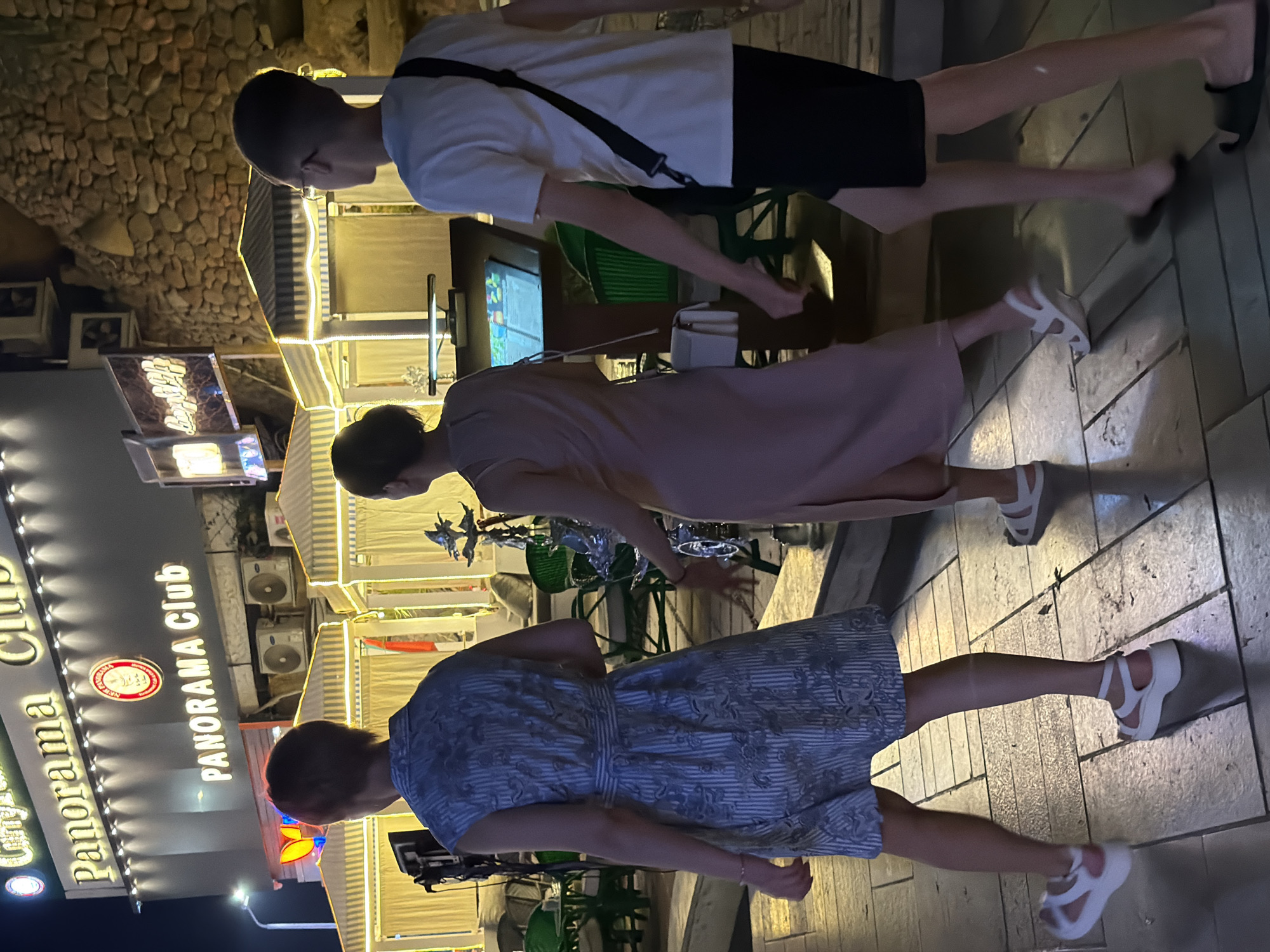 Three people, two women and one boy, walk along a tiled walkway at night. The woman on the left wears a sleeveless blue and white patterned dress and white platform sandals. The woman in the middle wears a pale pink sleeveless dress and white sandals. The boy on the right wears a white t-shirt, black shorts, and black slides. He carries a black strap across his chest.
Behind them, a lit building with the sign "Panorama Club" is visible. Several outdoor seating areas with striped awnings are illuminated with yellow lights. The background features a stone wall and greenery. A street lamp is also visible to the left. The scene appears to be in Sharm El Sheikh, Egypt, likely a tourist area.