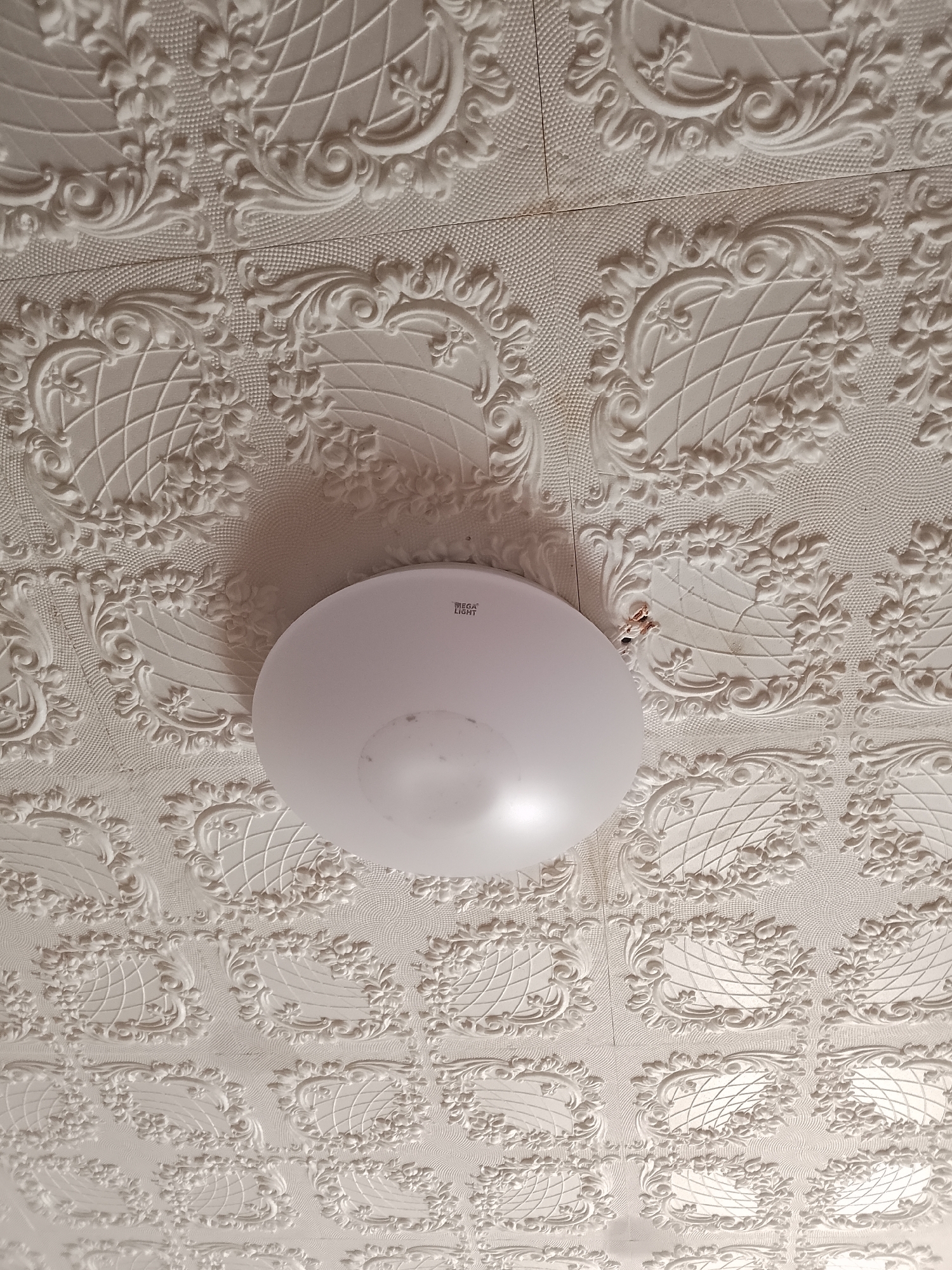 The image shows a white, embossed ceiling with a repeating floral and scroll pattern. A circular, white light fixture is centered on the ceiling. The lighting suggests the image was taken indoors, likely in a residential or public building. There is no visible information within the image to ascertain its location in the city of Abay, Kazakhstan.