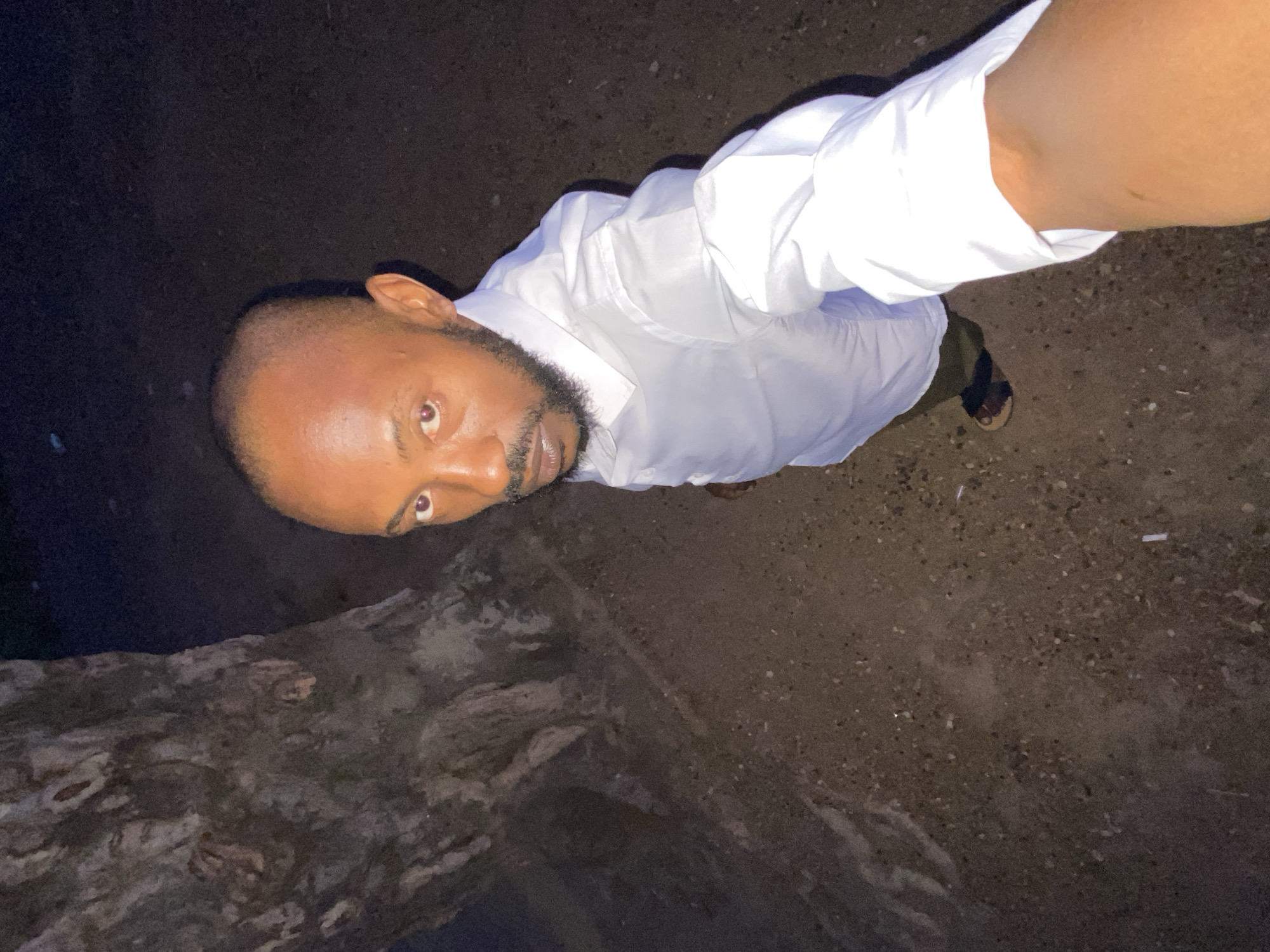 The image is a selfie taken from a high angle, looking down at a man in Sabon Tasha, Nigeria. The man has dark skin, a shaved head with a trimmed beard, and is wearing a white, button-down shirt. He is partially visible, appearing to be standing on rough ground or a poorly paved surface. To the left is a dark, textured area, possibly a wall or a large rock. The background is generally dark, indicating a time of low light. The visible shoe is a sandal. The man is the primary subject, with the environment contributing to the context.