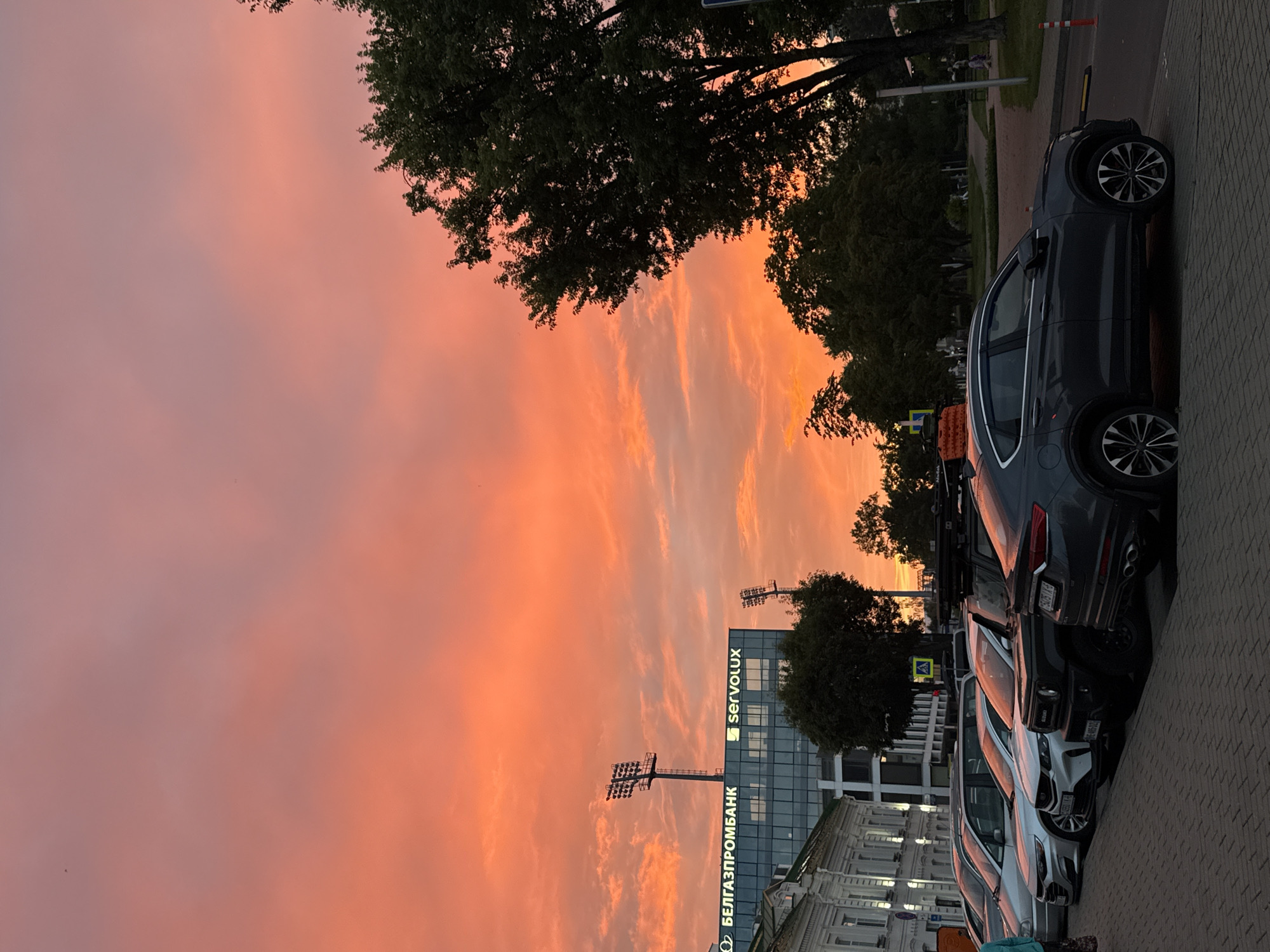A panoramic view of a city street in Mahilyow, Belarus, captures a dramatic sunset painting the sky in vibrant shades of orange and pink. In the foreground, a row of parked cars lines a brick-paved street. Among them are a dark grey SUV with silver rims, a white SUV, and a silver minivan. Behind the vehicles, a modern glass-fronted building stands tall, bearing the logos "БЕЛГАЗПРОМБАНК" and "servolux." Two tall light towers are visible on top of the building. To the right, lush green trees frame the scene, their leaves silhouetted against the colorful sky. A few distant figures can be seen walking in a park-like area to the right, adding a sense of everyday life to the picturesque setting. The overall impression is one of urban tranquility bathed in the warm glow of a setting sun.
