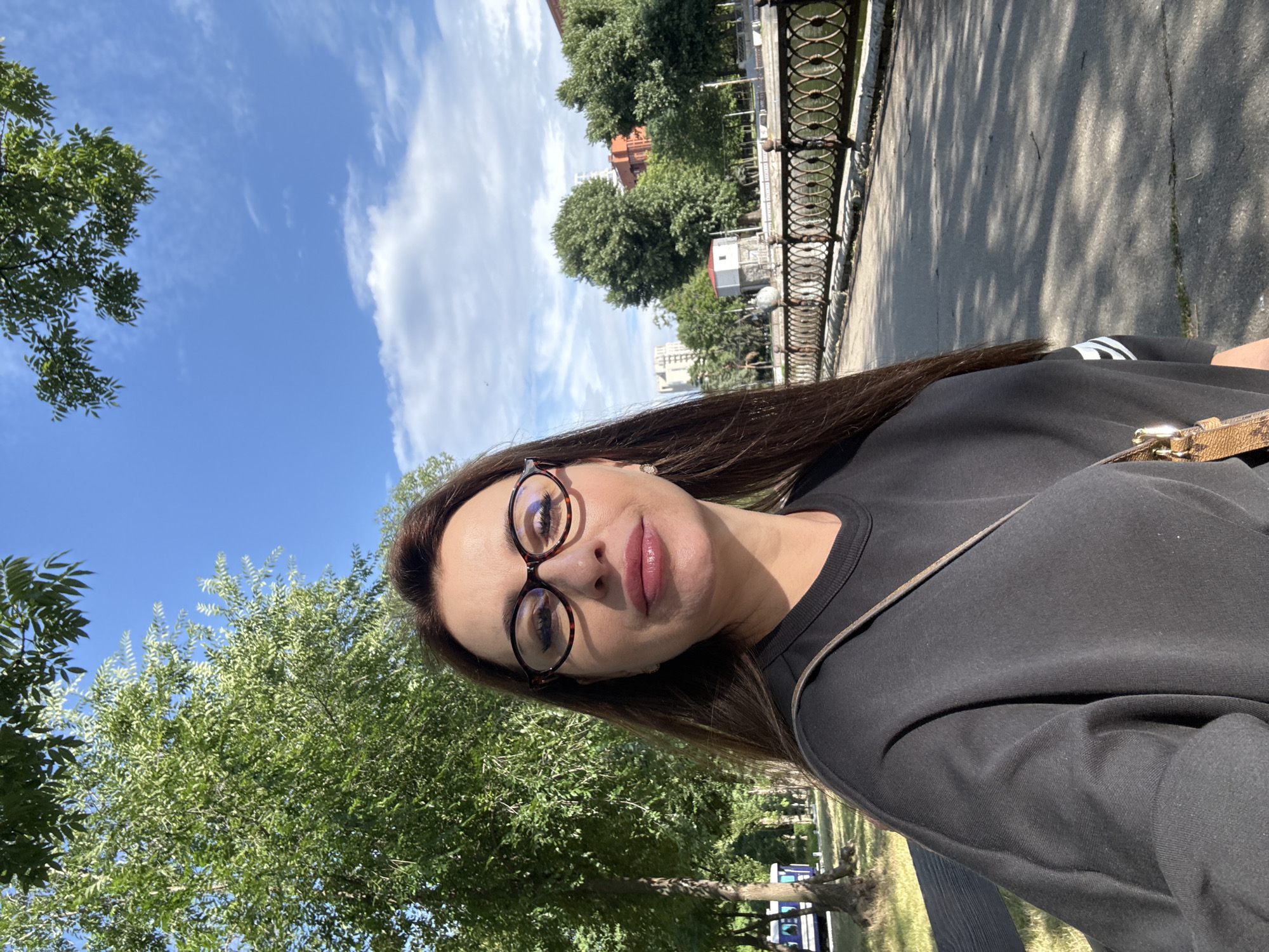 In the city of Dnipro, Ukraine, on a bright, sunny day, a woman with long, dark brown hair and glasses takes a selfie in a park. She is wearing a black sweatshirt and a light-colored crossbody bag. The background shows lush green trees, a blue sky with white clouds, and a paved walkway with a decorative metal fence along a body of water. Buildings and more trees are visible in the distance.