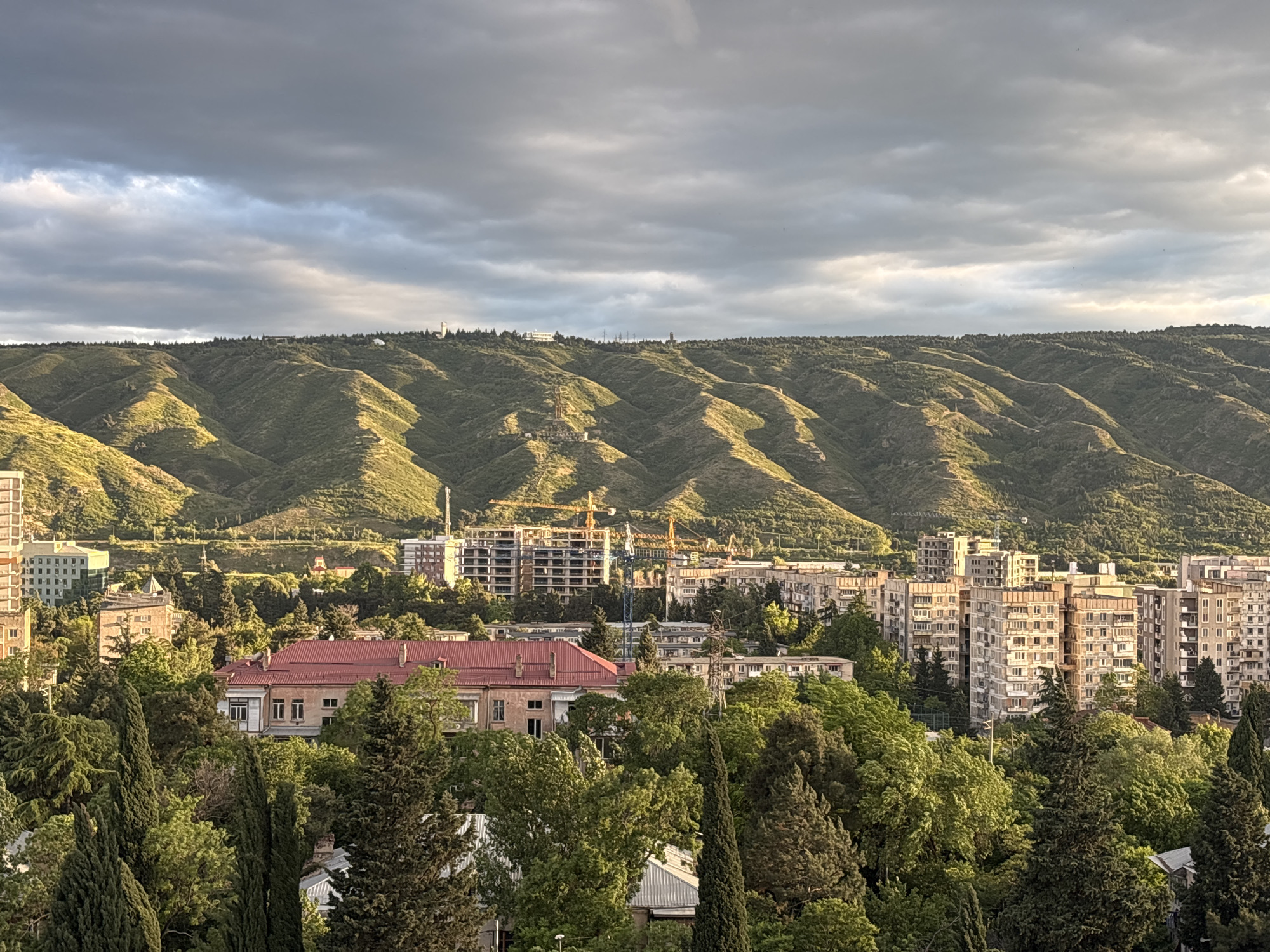This image shows a cityscape in Tbilisi, Georgia, under a cloudy sky. The foreground features dense green trees, interspersed with residential buildings. Behind them, mid-rise apartment buildings and a structure under construction with cranes are visible. In the background, a series of green hills or mountains stretch across the horizon. The light is somewhat diffused, indicating an overcast or late afternoon/early morning setting. The built environment is partially obscured by greenery, suggesting a city with a significant amount of vegetation.
