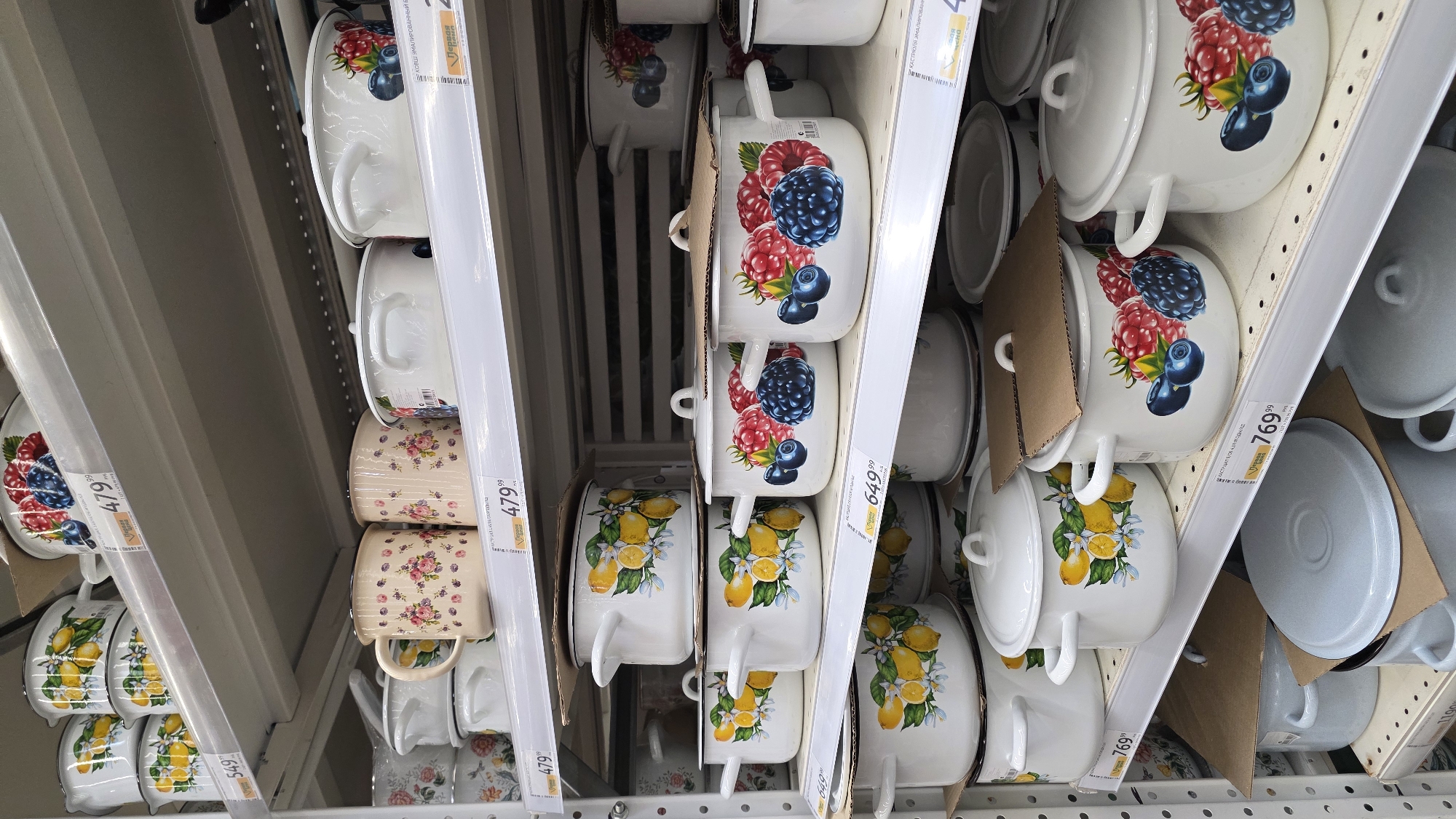 The image captures a retail store aisle, specifically a section displaying various enamel pots and pans on metal shelving units. The cookware is stacked and arranged neatly, with some items still in cardboard packaging or wrapped in plastic.
The primary subjects are diverse enamel pots and mugs, showcasing different designs. Many pots are white with vibrant illustrations: one prominent design features a cluster of red raspberries and dark blue blueberries, while another displays bright yellow lemons with green leaves and white blossoms. A different set of smaller pots or mugs, visible on the left and right, features a delicate, light-colored floral pattern, possibly pink or purple flowers on a cream background. Alongside these decorative items, there are plain grey enamel pots and lids, suggesting a more basic or utilitarian range.
The scene depicts a typical merchandising setup in a supermarket or home goods store. There are no people visible, suggesting the photo might have been taken during less busy hours or focusing solely on the products. The items are displayed for sale, with multiple price tags attached to the shelves.
Notable details include the various price tags. Visible prices include "479.99", "549.99", "649.99", and "769.99". The text on these tags and product labels is in Russian. Specifically, "КАСТРЮЛЯ ЭМАЛИРОВАНН" translates to "Enameled Pot," and "КОВШ ЭМАЛИРОВАННЫЙ" means "Enameled Saucepan" or "Ladle." Another readable phrase is "Первая Цена," which translates to "First Price" or "Best Price," commonly used in retail. The store lighting is bright and even, consistent with an indoor retail environment.