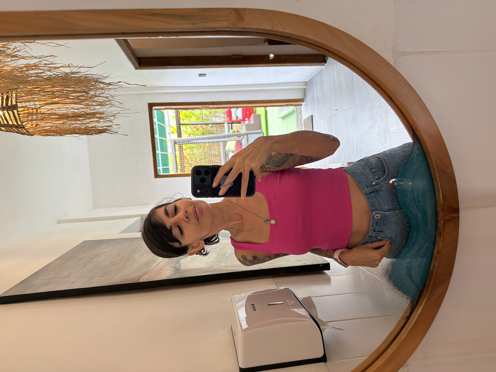 A woman with short, dark hair is taking a mirror selfie in a brightly lit bathroom, likely in Kuta Selatan, Indonesia. She is wearing a bright pink, ribbed tank top and blue denim jeans, with a silver necklace around her neck. Her left arm, adorned with tattoos, is raised to hold her phone, and her right hand is in her jeans pocket. The bathroom features white tiled walls and a wooden-framed oval mirror. To her left, a white paper towel dispenser is mounted on the wall. The reflection in the mirror reveals an open doorway leading to an outdoor area with lush greenery and some red and yellow seating. A woven pendant light fixture hangs from the ceiling. The floor appears to be tiled with light-colored square tiles.