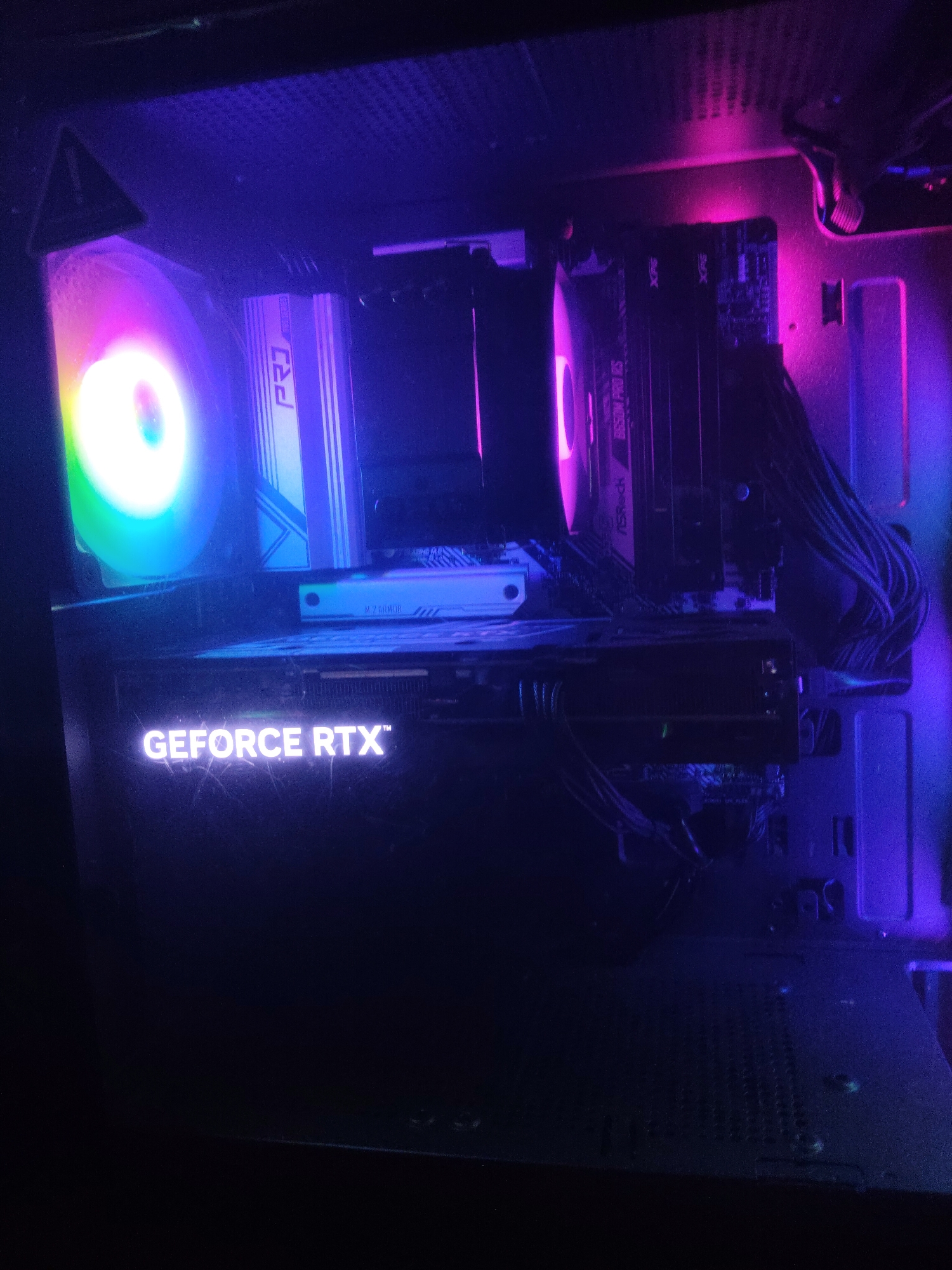 A dimly lit view inside a computer case. Visible components include a GeForce RTX graphics card, RAM modules, CPU cooler, and motherboard with M.2 SSD. The scene has blue and purple lighting. The location in the city of Perm, Russia, is not discernible.