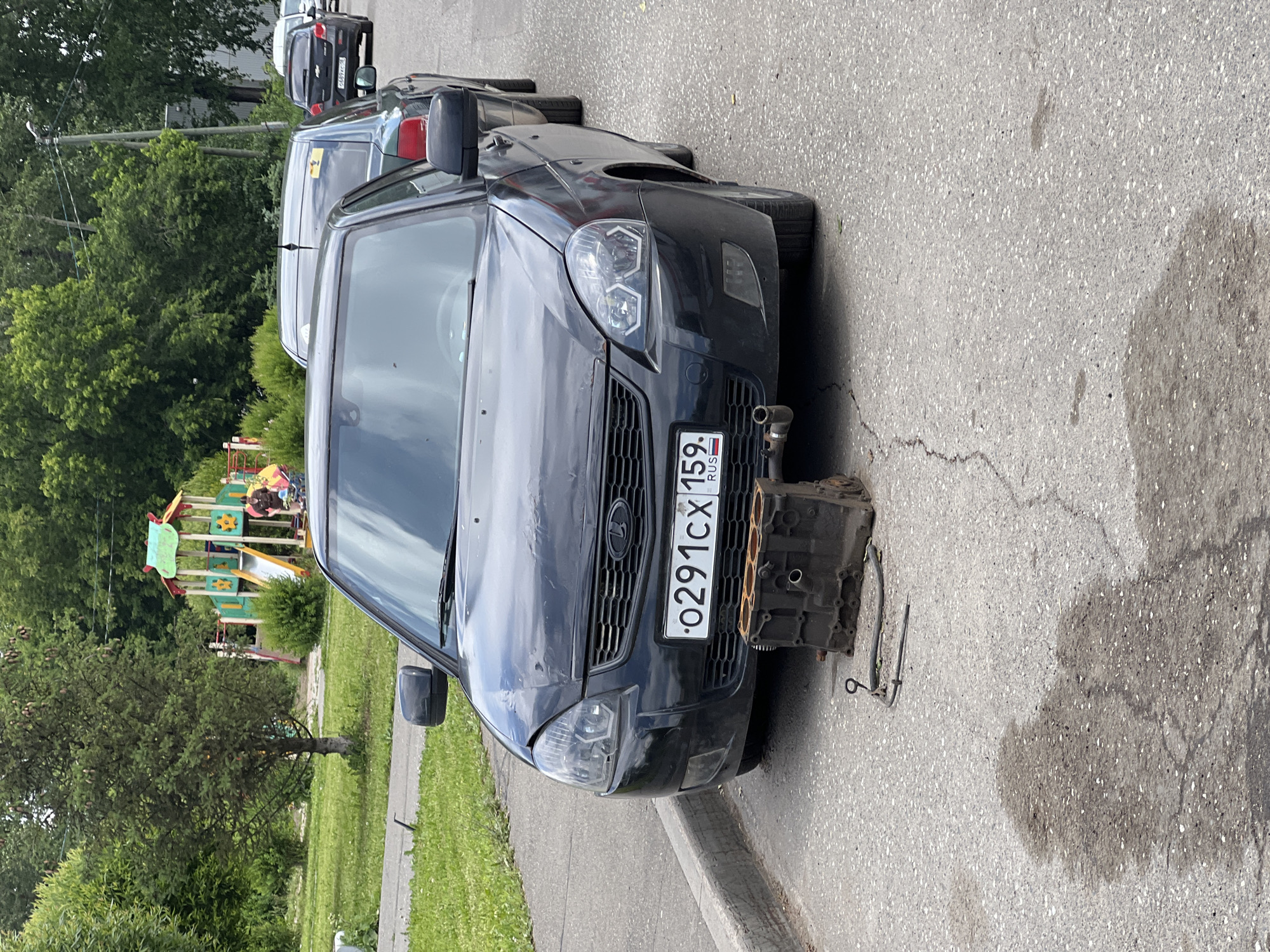 In the city of Izhevsk, Russia, on a paved road next to a sidewalk, a black sedan is parked with its hood slightly ajar. In front of the vehicle, resting on the pavement, is a disassembled engine block. Nearby, a dipstick and a metal tube are also visible on the ground. The background features a playground with slides and climbing structures, surrounded by green trees and grass. Other cars are parked in a line behind the primary vehicle. A dark liquid stain is present on the pavement in front of the engine block.